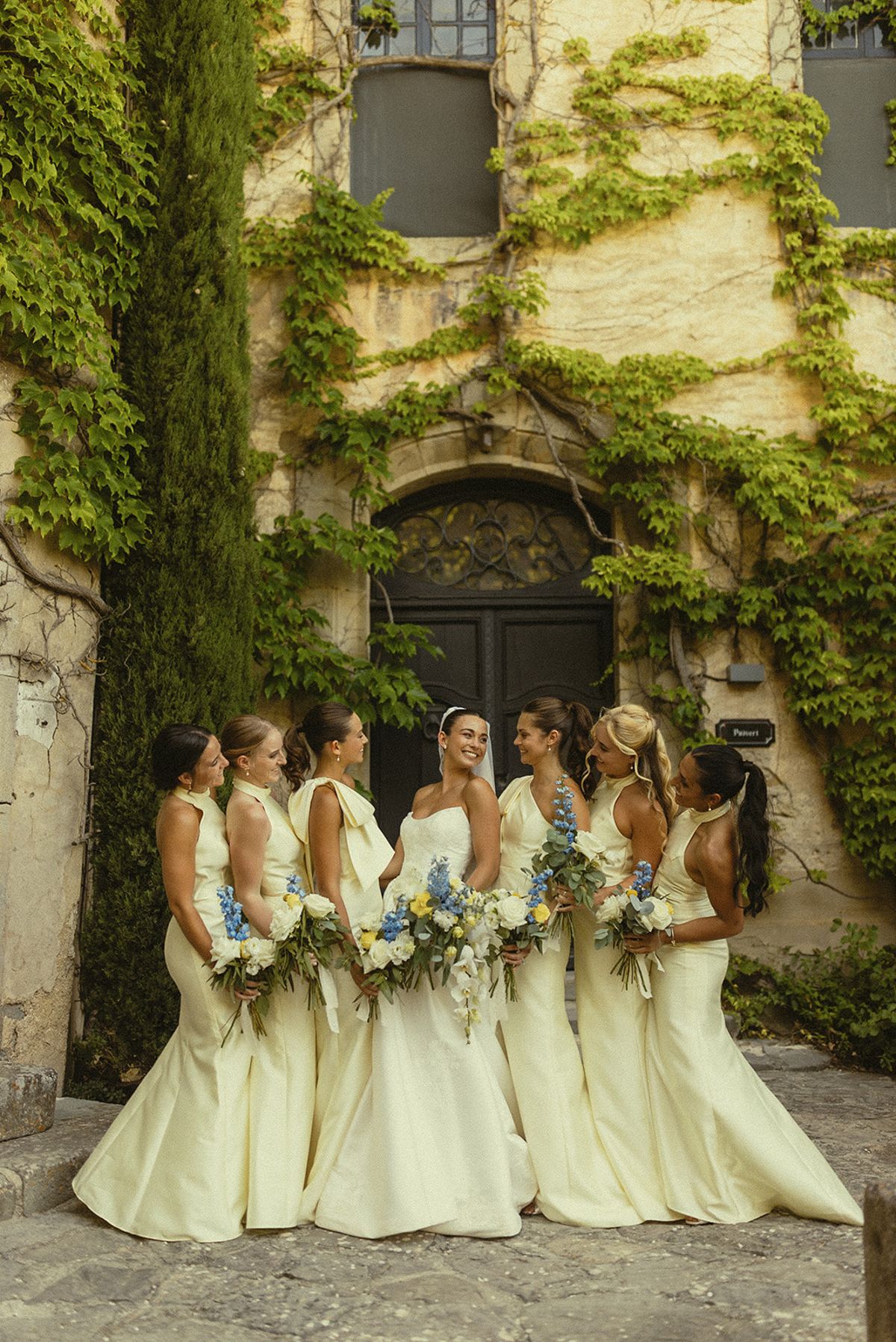 Seven bridesmaids in pale yellow gowns stand close together holding bouquets, facing an ivy-covered stone wall and dark door.