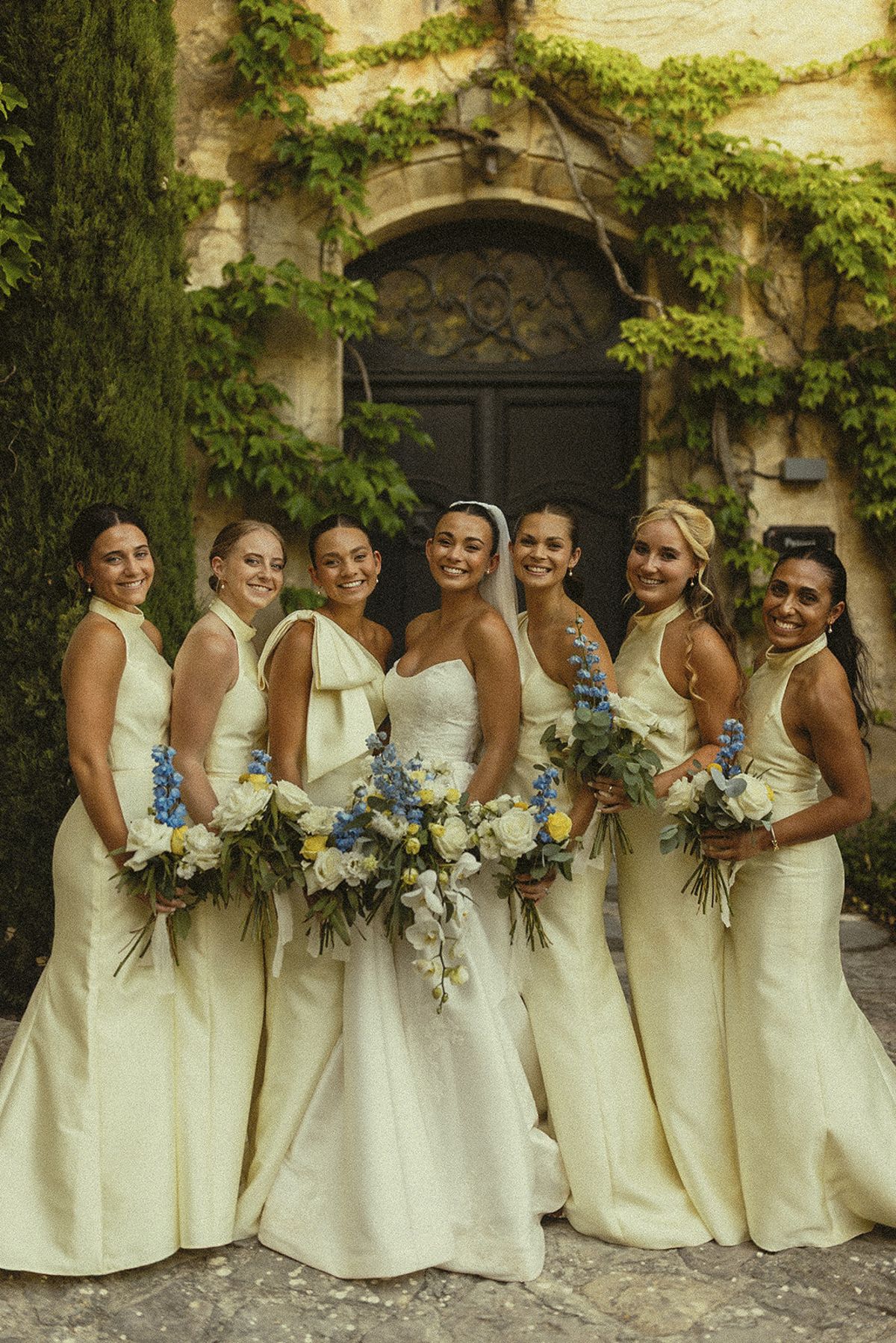 A group of bridesmaids in cream gowns stand close together holding blue and white bouquets in front of an ivy-covered doorway.