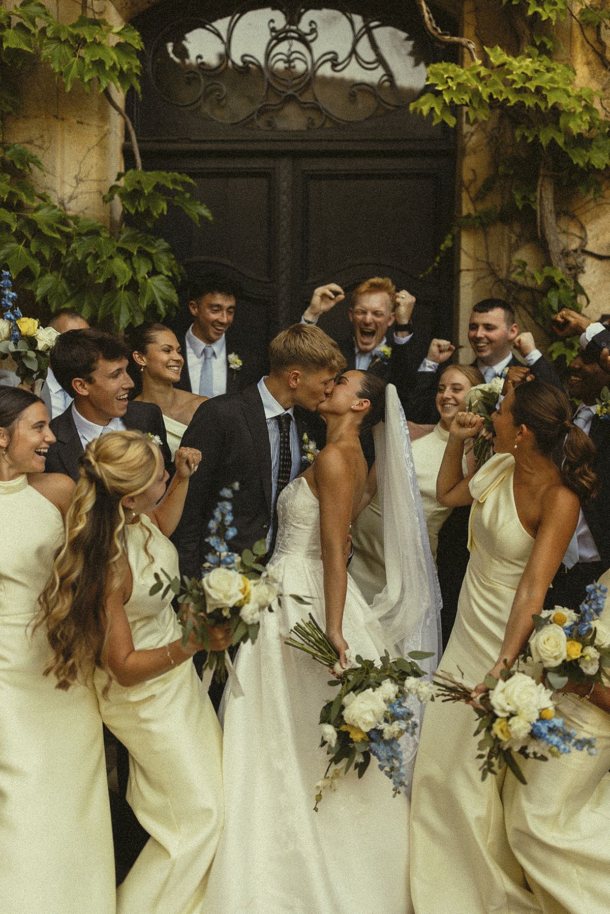The couple kiss while bridesmaids holding bouquets and groomsmen cheer closely around them in front of a large dark door.