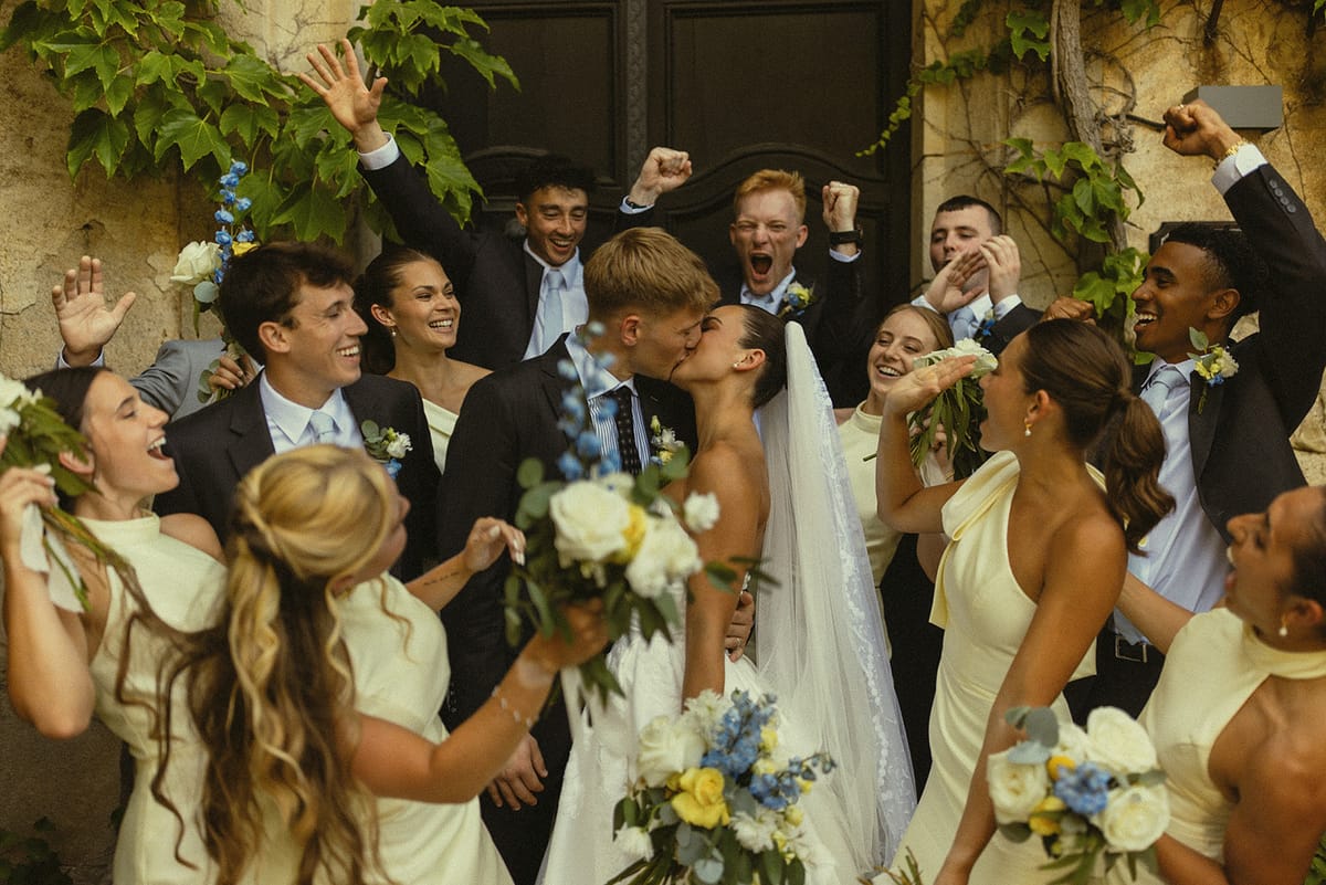 Bridesmaids and groomsmen cheer with raised arms around the couple as they kiss, holding blue and white bouquets.