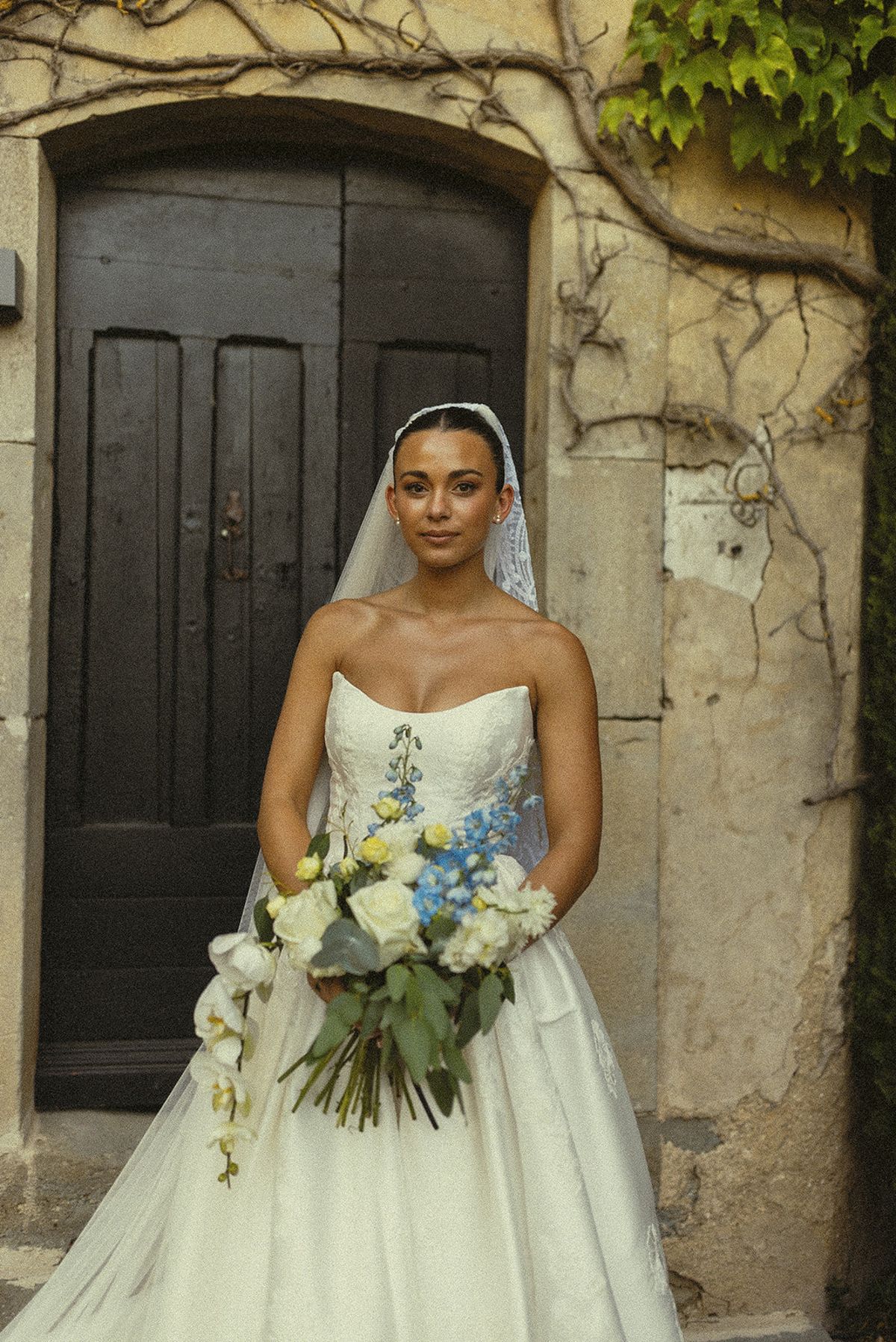 A bride in a strapless white gown and veil holds a bouquet of blue and white flowers in front of an old wooden door.