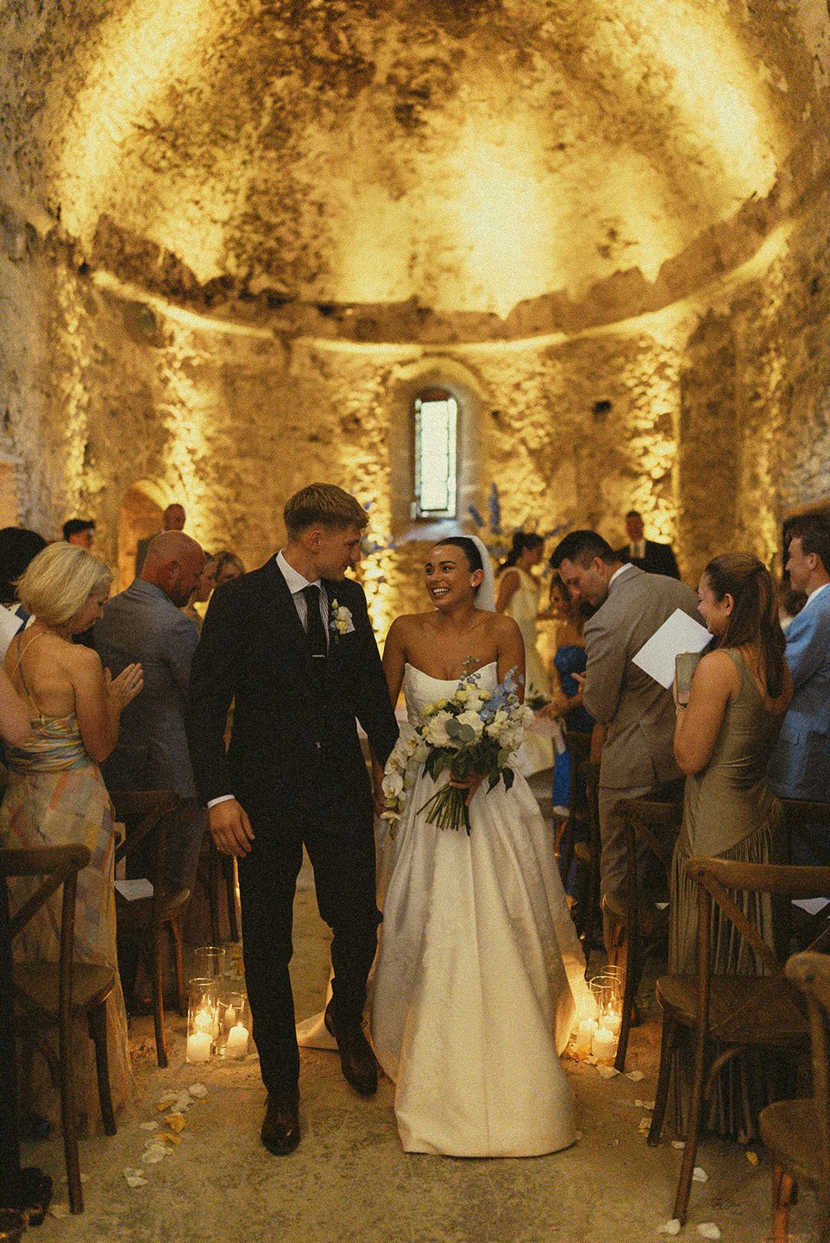 The couple walk smiling down the aisle of a candlelit stone chapel between rows of seated guests.