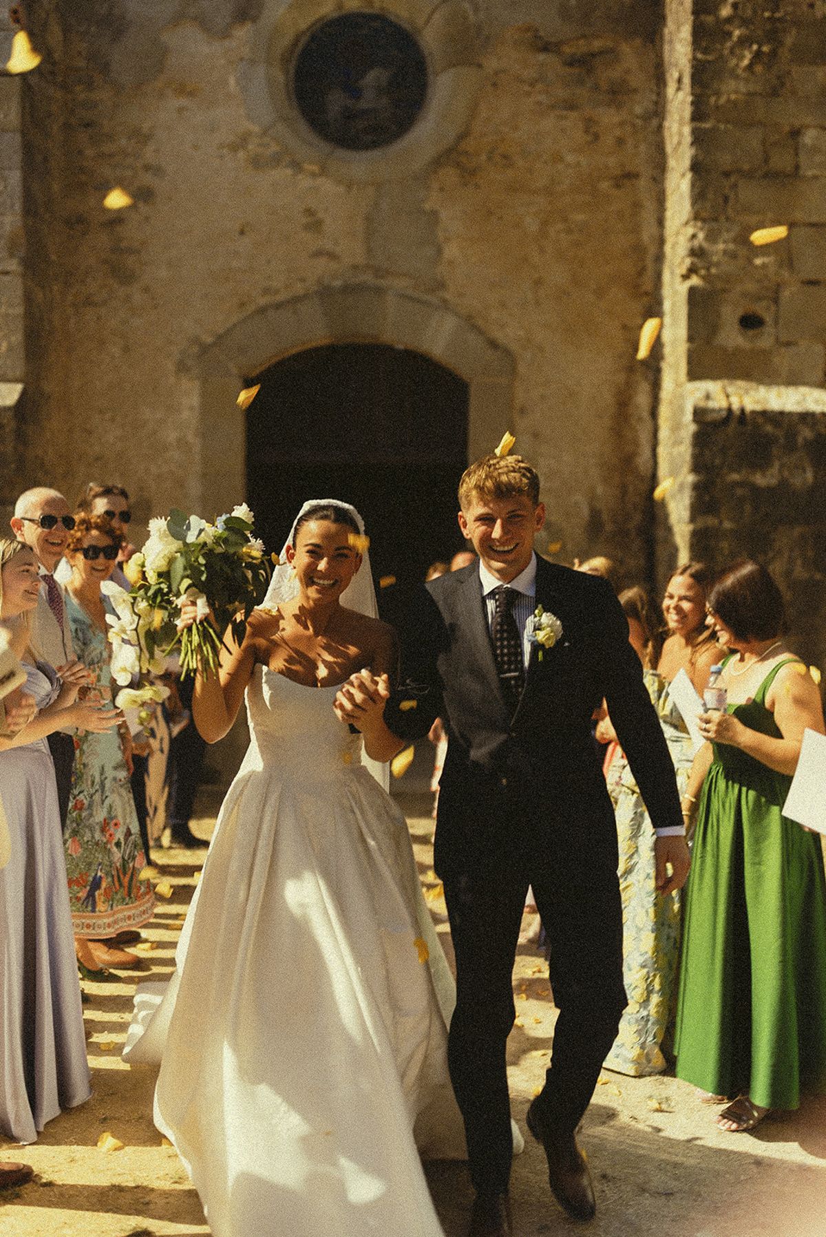 The smiling couple walk out together past guests who throw confetti, in front of an old stone building.