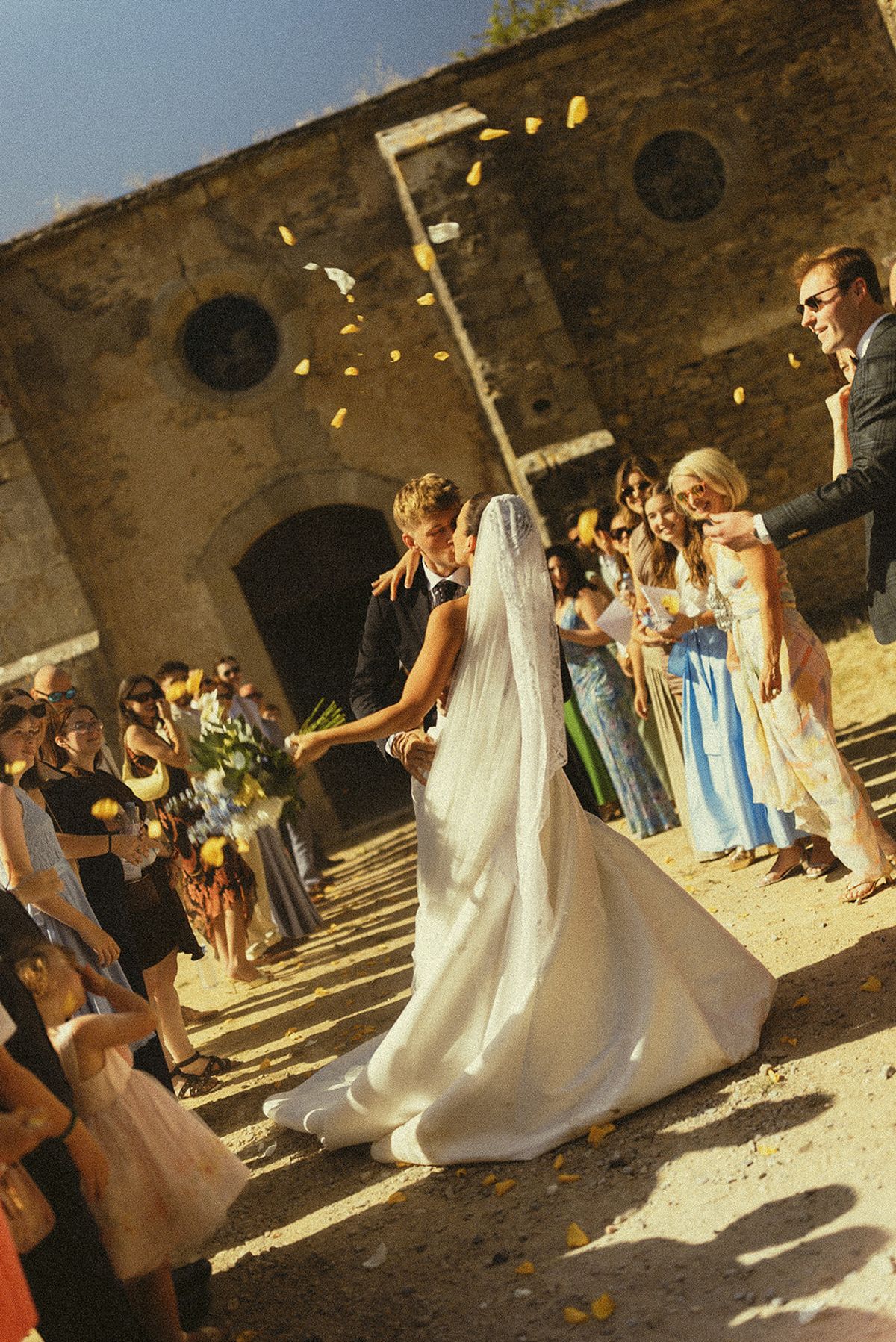 The bride in a long veil walks between guests who throw petals into the air beside an old stone building.