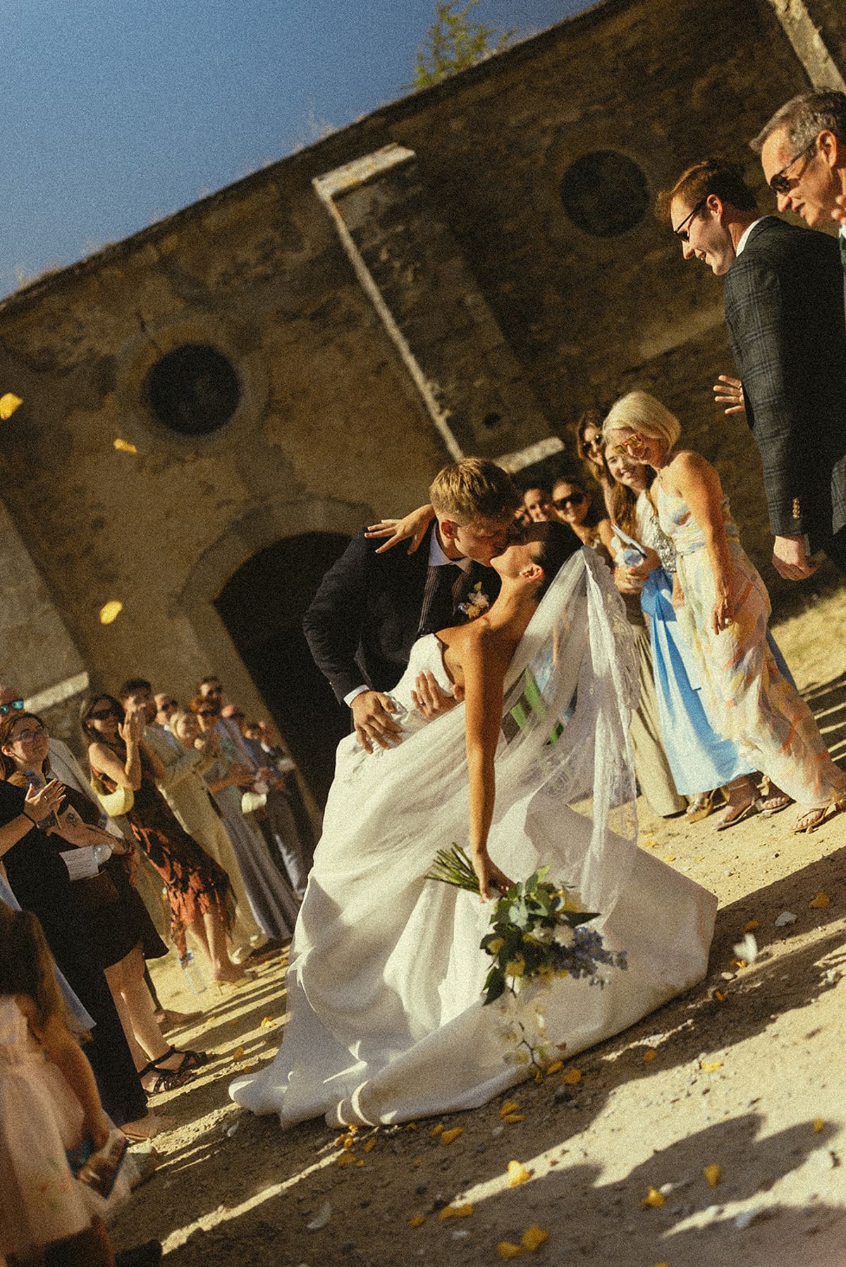 The couple pass through a courtyard as guests cheer and throw confetti, the bride's veil and gown billowing around her.