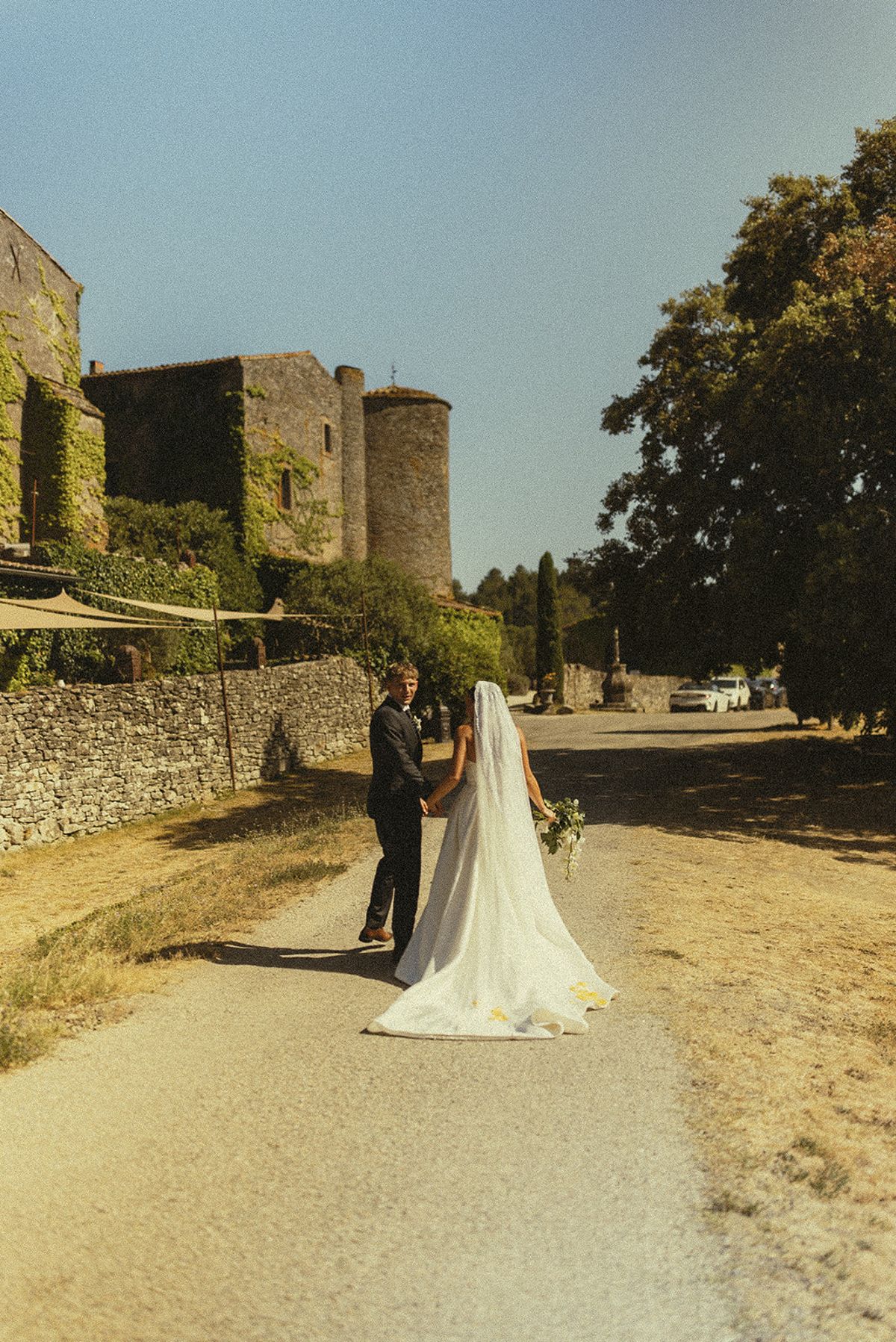 The couple stand together on a gravel path, seen from behind, with the bride's long veil trailing and a stone castle beside them.