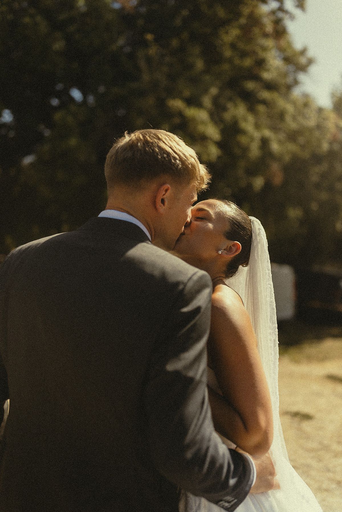 The groom in a grey suit kisses the bride, who wears a long veil, in warm sunlight.