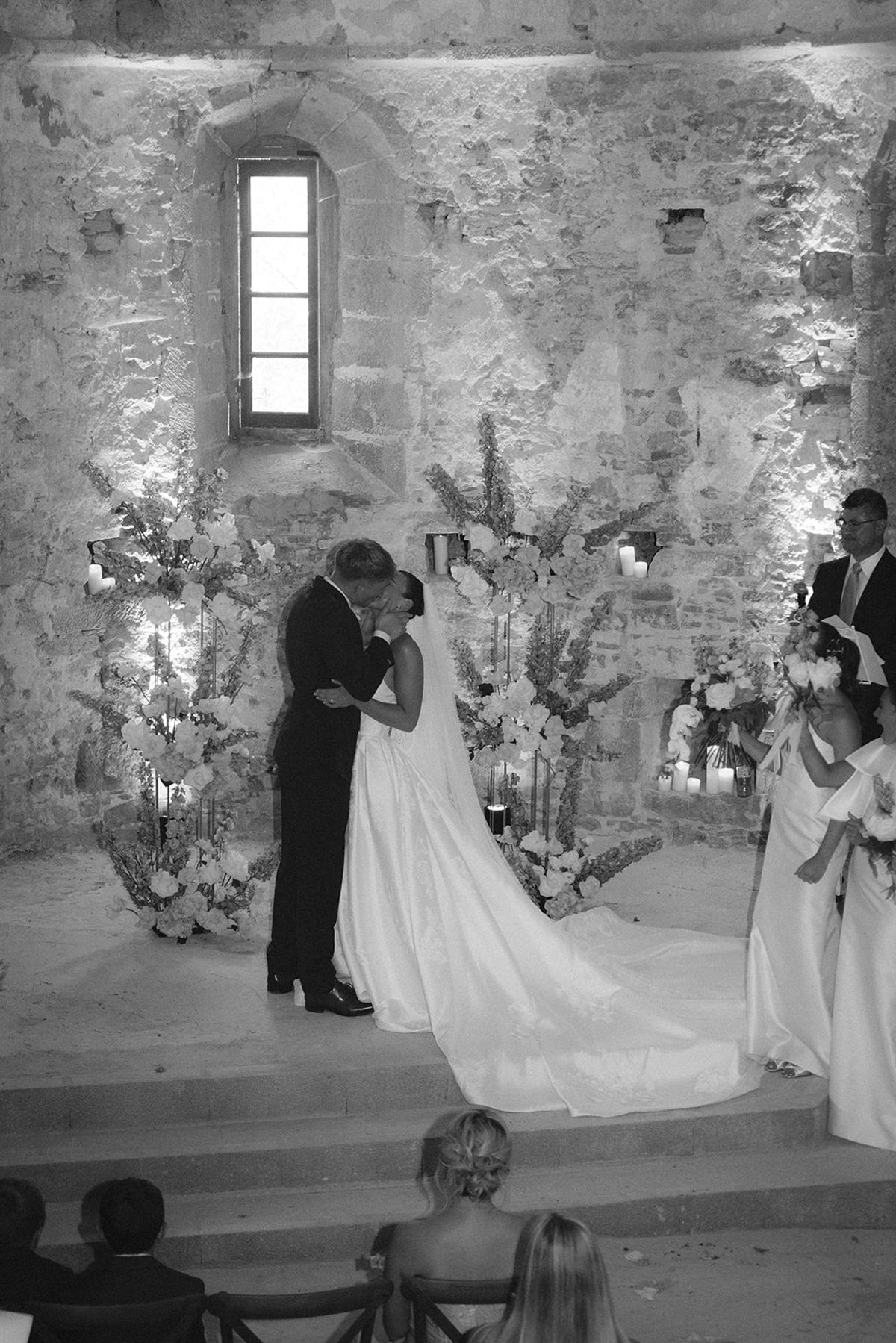 In black and white, the couple kiss at an altar decorated with flowers while bridesmaids stand beside them and guests watch.