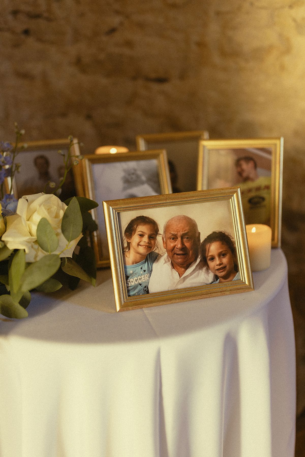 Gold-framed photographs stand on a table with a white cloth, next to a candle and white flowers.