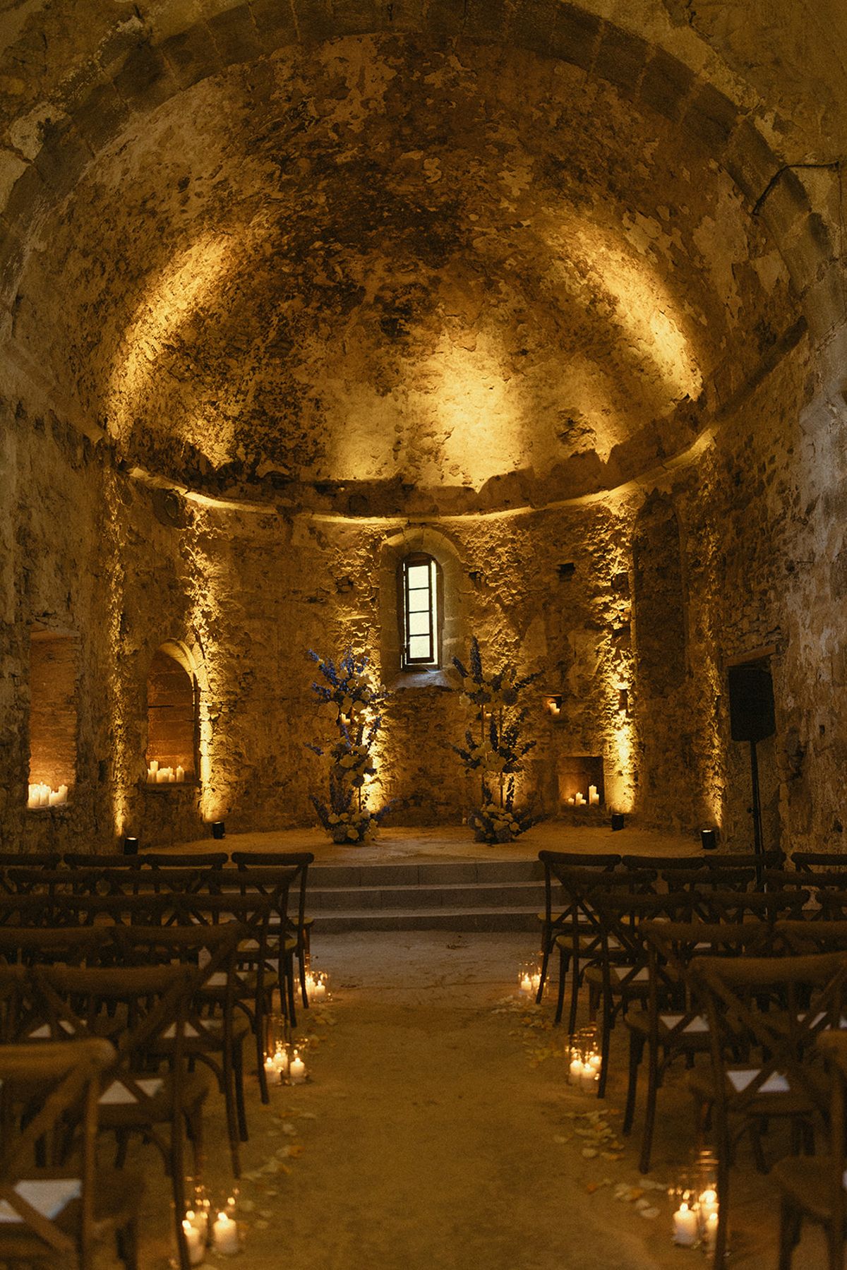 A vaulted stone chapel lit in warm tones, with rows of wooden chairs and candles along the central aisle.