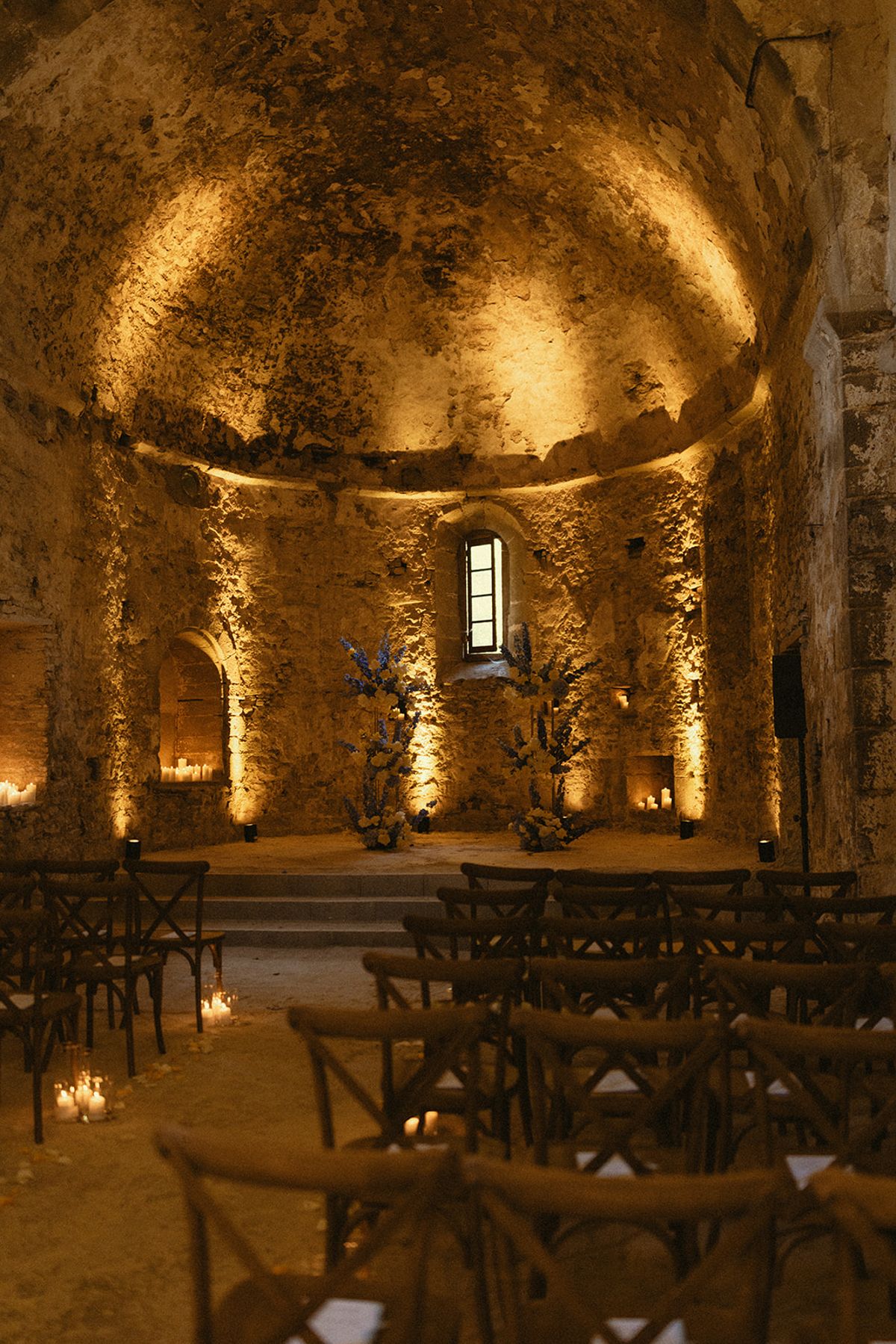 A vaulted stone chapel glows with warm lighting and candles, with rows of wooden chairs facing a small altar.
