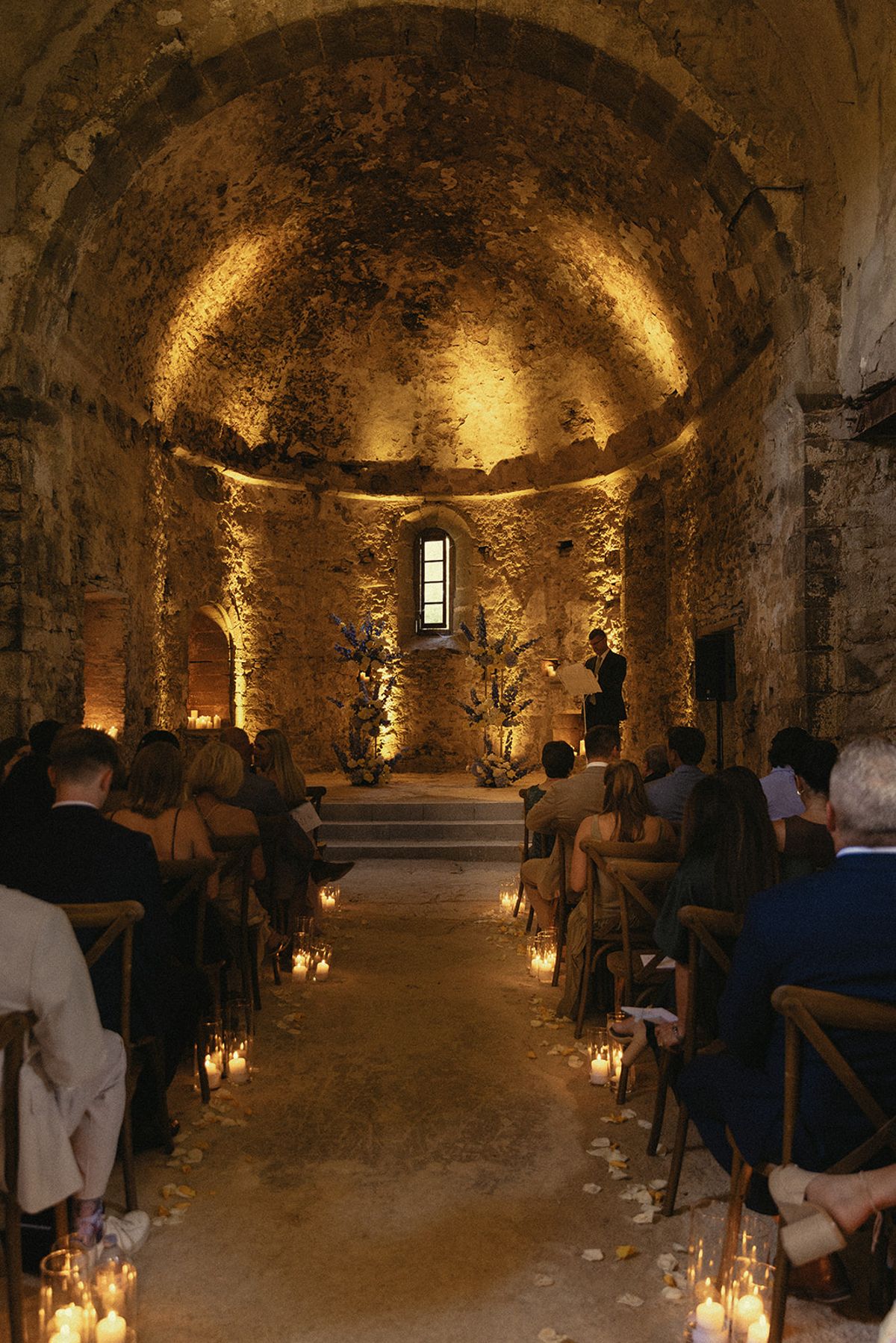 Guests sit on both sides of a candlelit aisle leading to a glowing, vaulted stone apse in a chapel.