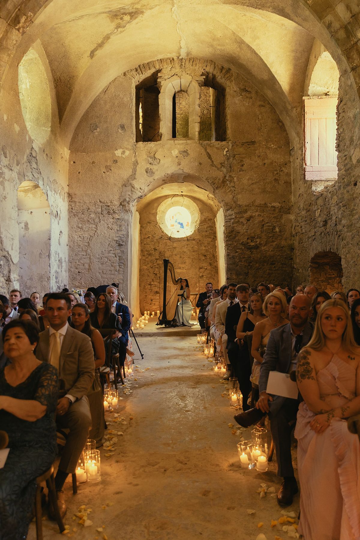Guests sit on both sides of a candlelit aisle inside a vaulted stone chapel, looking towards the altar.