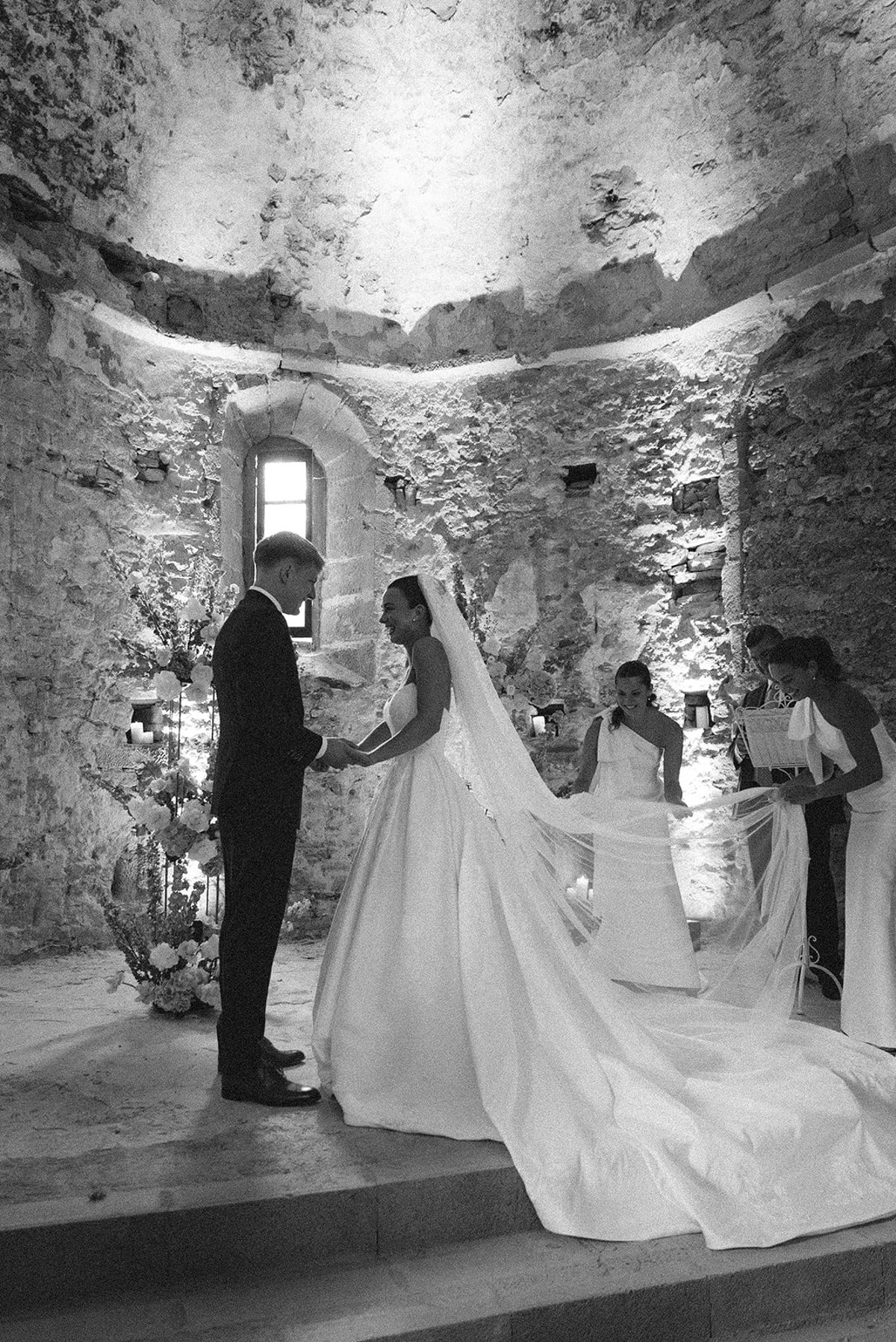 In black and white, the groom and the bride in a long train stand in a stone chapel apse with two women behind.