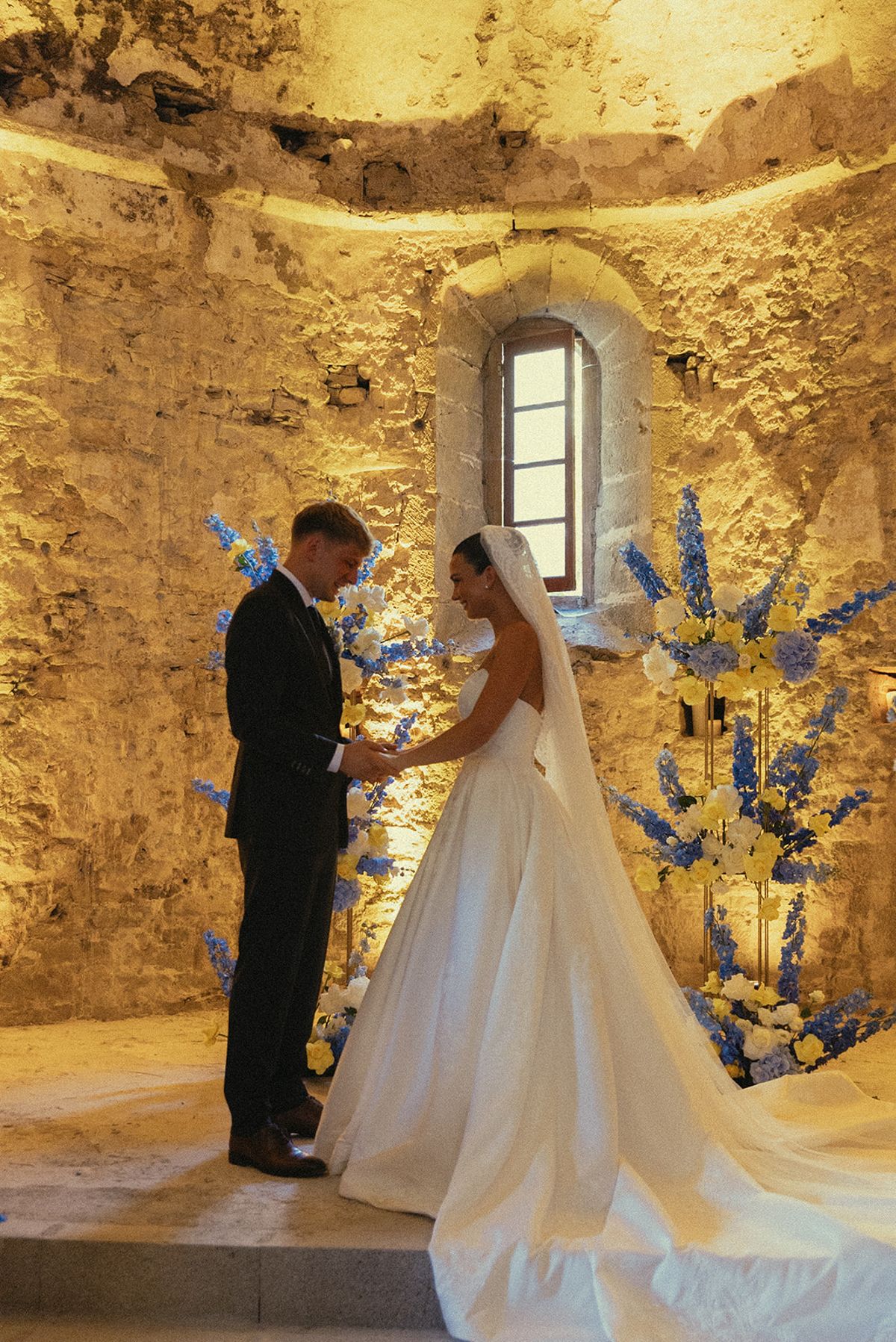 The groom in a dark suit and the bride in a long veil and gown face each other inside a lit stone chapel.