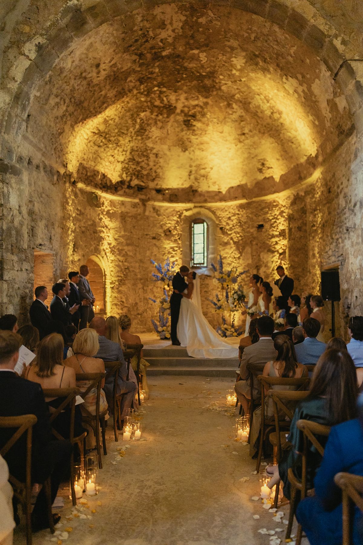 Guests sit on wooden chairs either side of a candlelit aisle as the couple stand at the altar of a glowing stone chapel.
