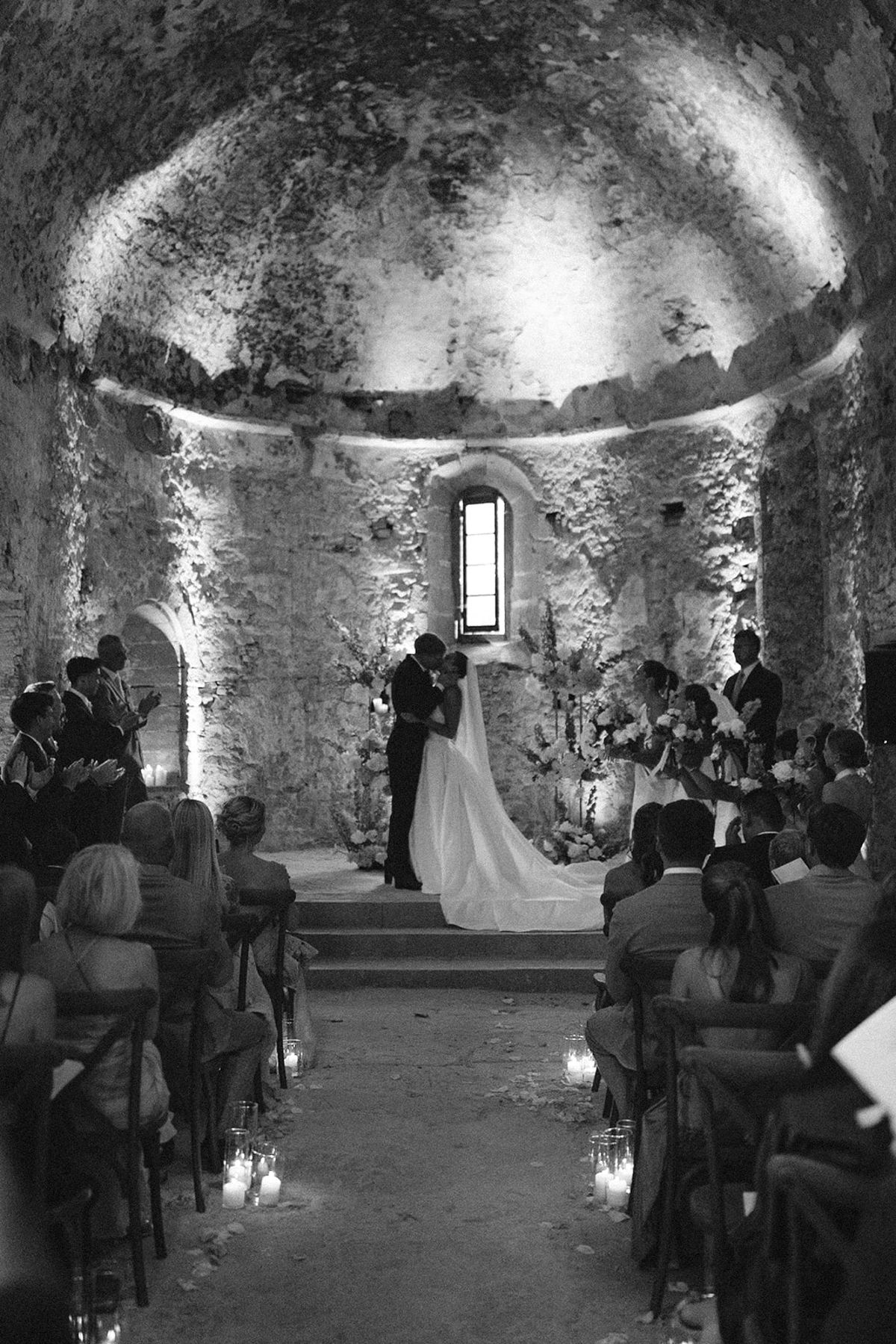 In black and white, guests sit along a candlelit aisle facing the couple at the altar of a vaulted stone chapel.