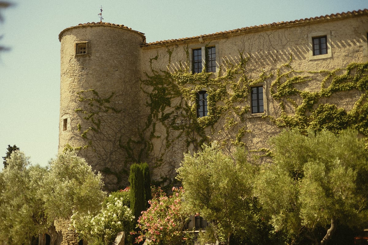 A round stone tower and long stone building with small windows rise above green shrubs under a blue sky.