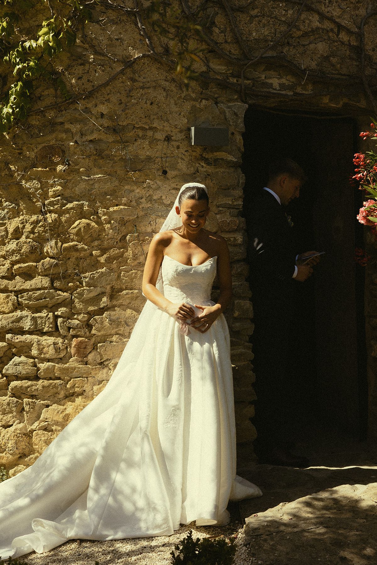 A bride in a strapless white gown stands against a sunlit stone wall while a man in a dark suit waits at a doorway.
