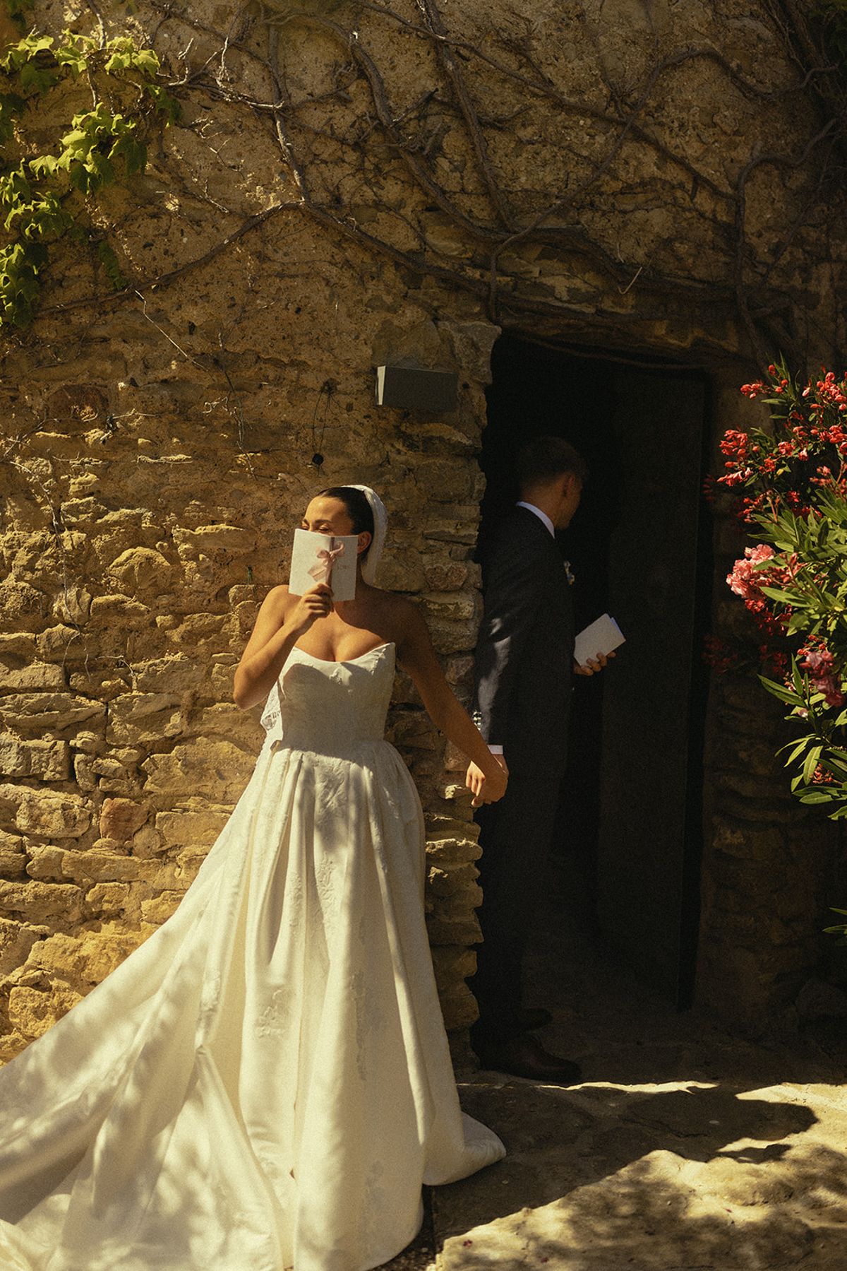 The bride in a white gown covers her face with one hand beside the groom, who stands in a dark stone doorway.