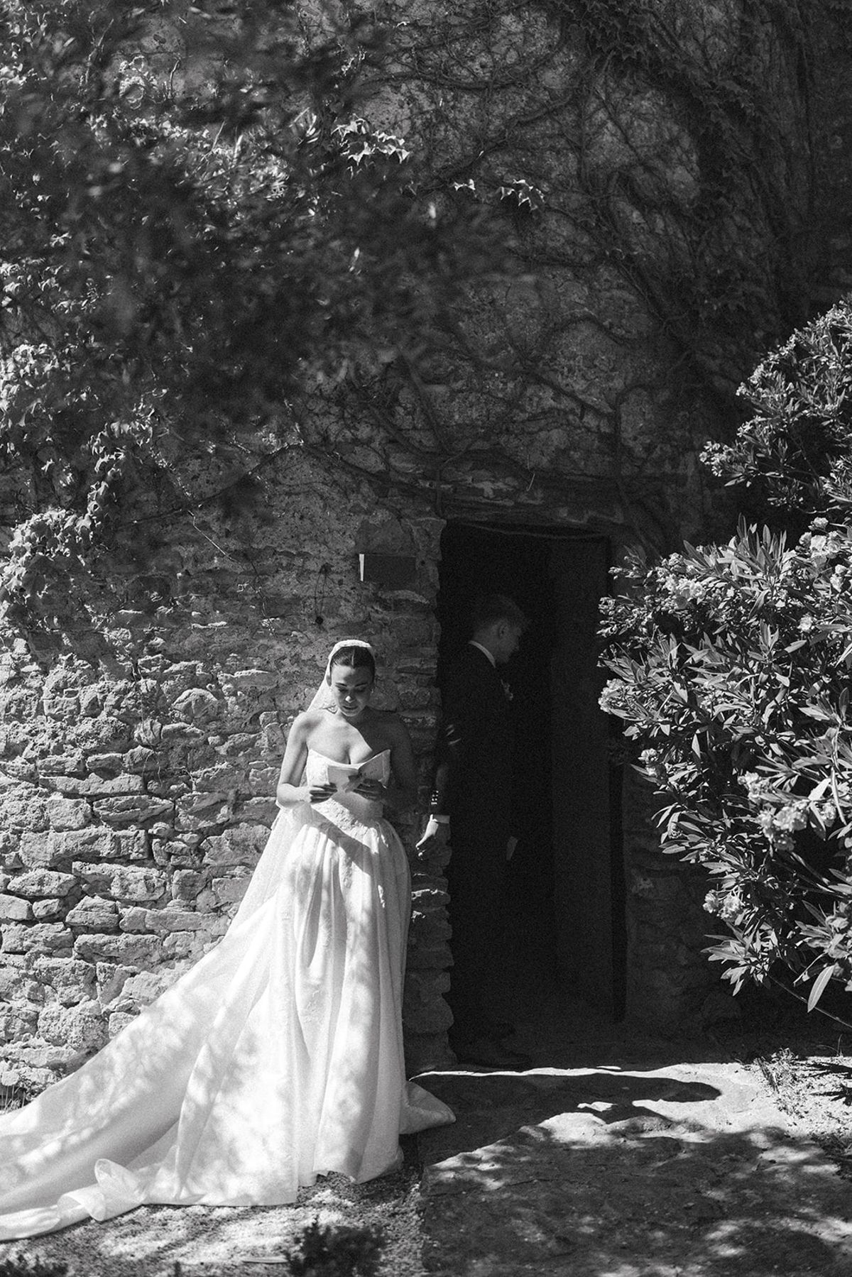 In black and white, a bride in a long veil stands by a stone wall and shrubs while a man waits in a doorway.