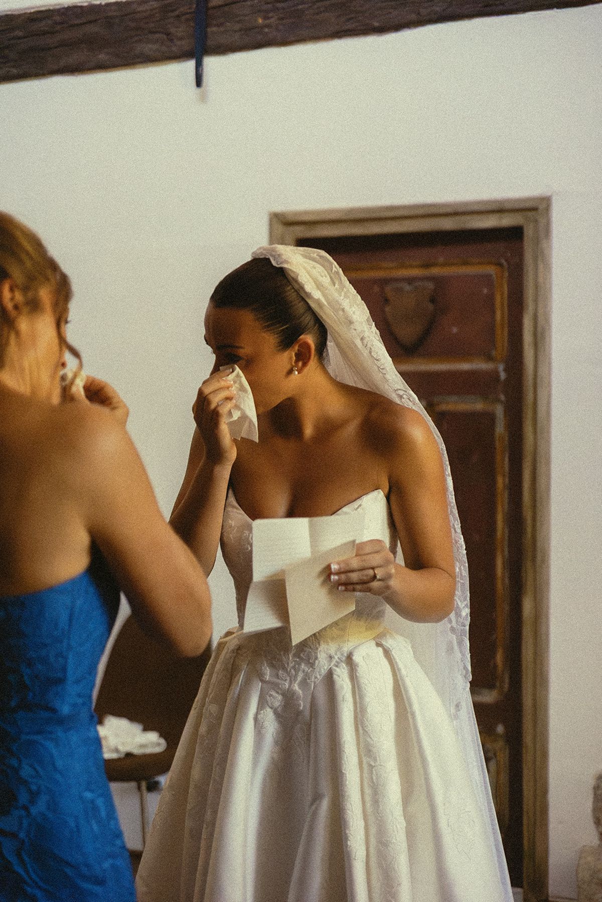 A bride in a veil dabs her eyes with a tissue and holds a card, standing beside a woman in a blue dress.