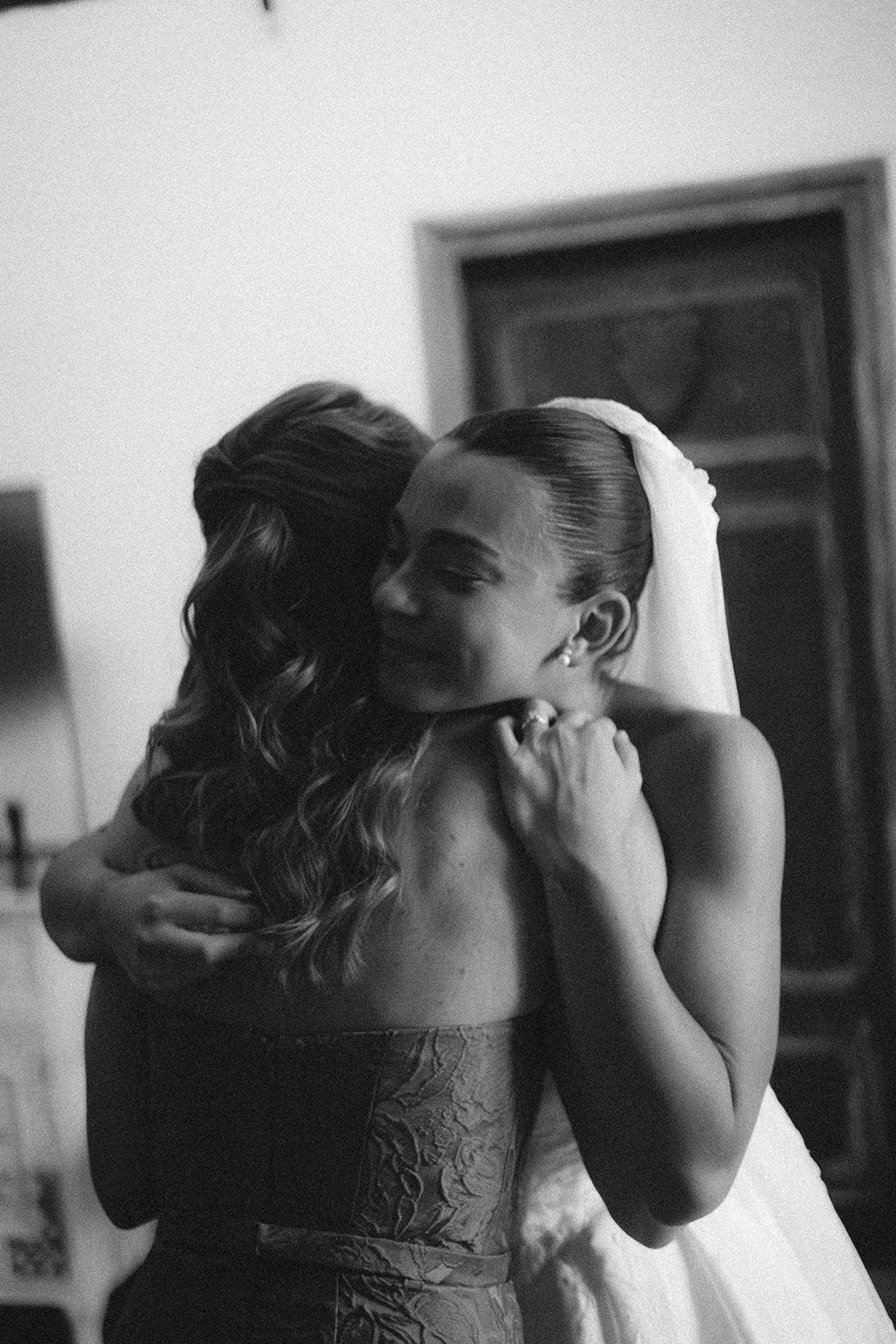 In black and white, a smiling bride in a veil hugs a woman with long wavy hair beside a wooden door.