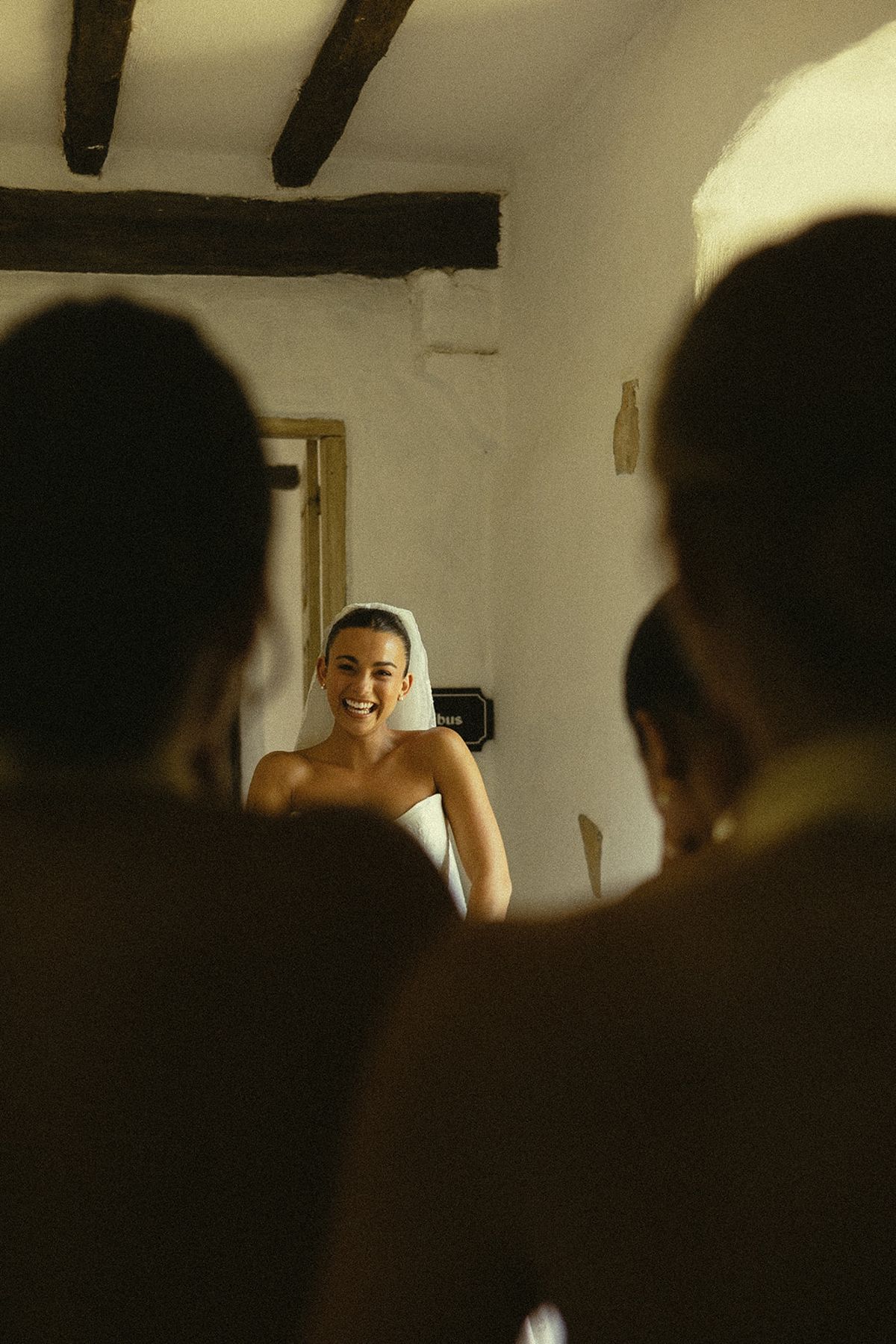A smiling bride wearing a veil stands in a whitewashed room, framed by the dark silhouettes of people in the foreground.
