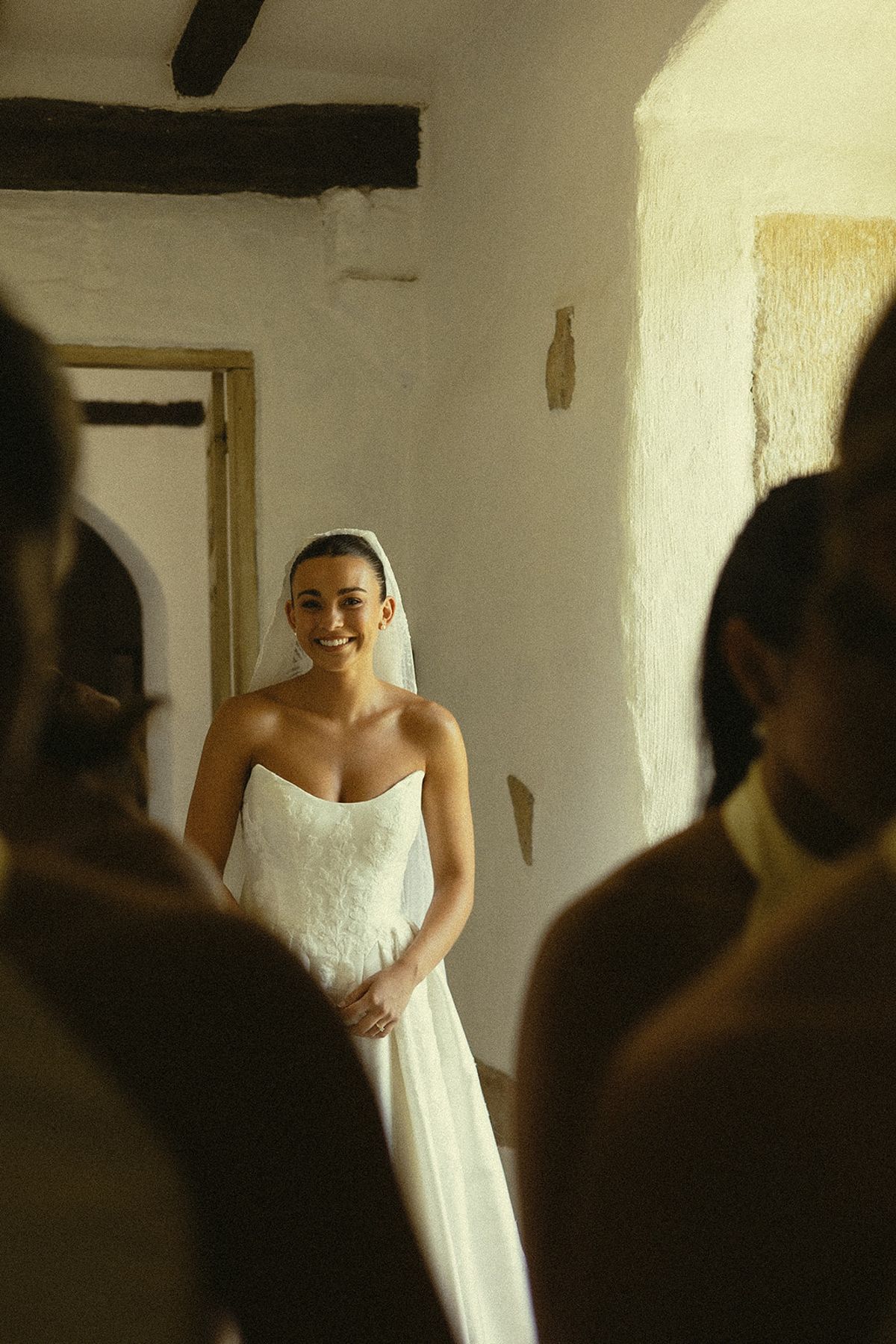The bride in a strapless white gown and veil smiles warmly while standing in a stone passage, framed by dark silhouettes of guests in the foreground.