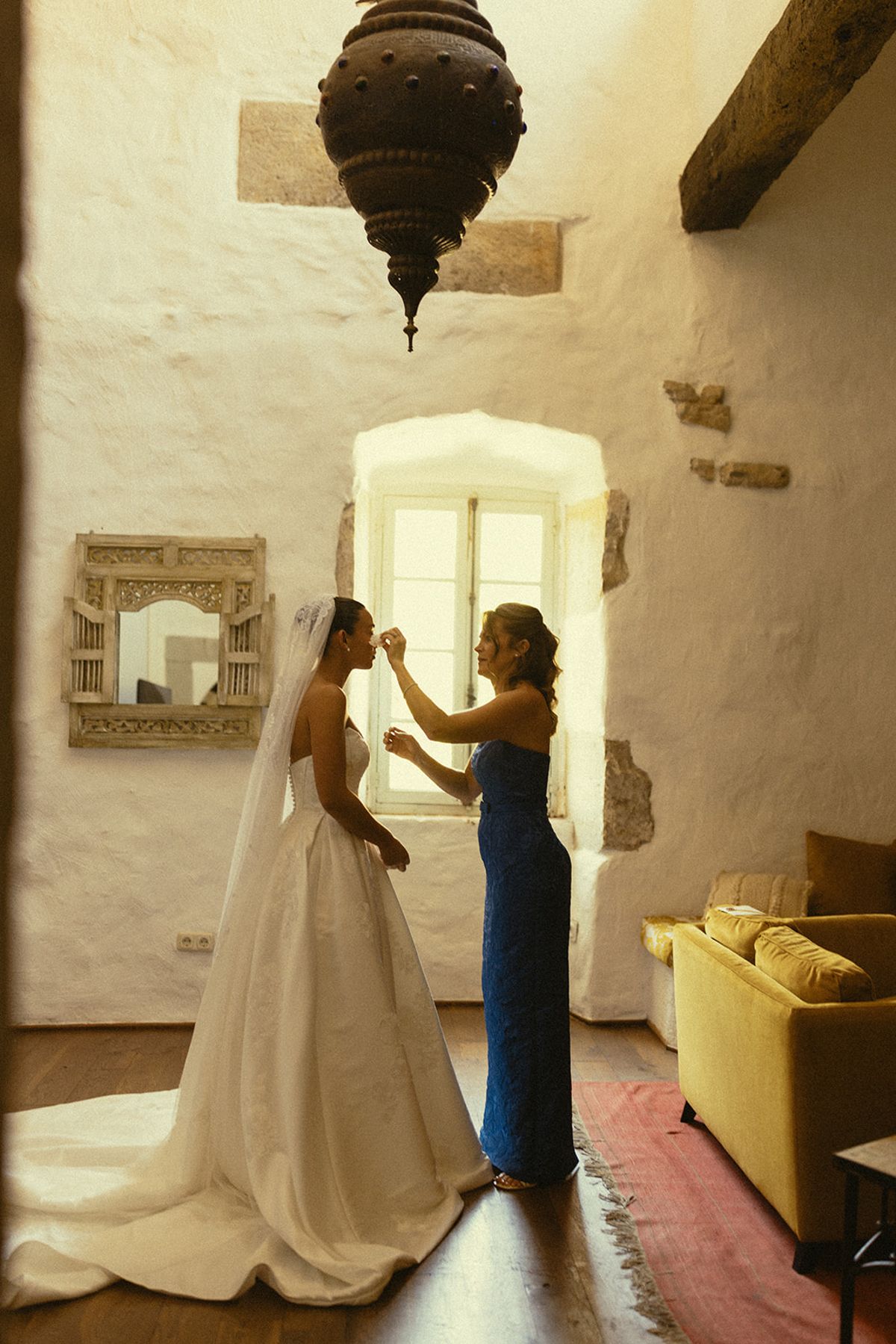 The bride in a long white gown stands by a tall window with a woman in a blue dress who reaches towards her veil.