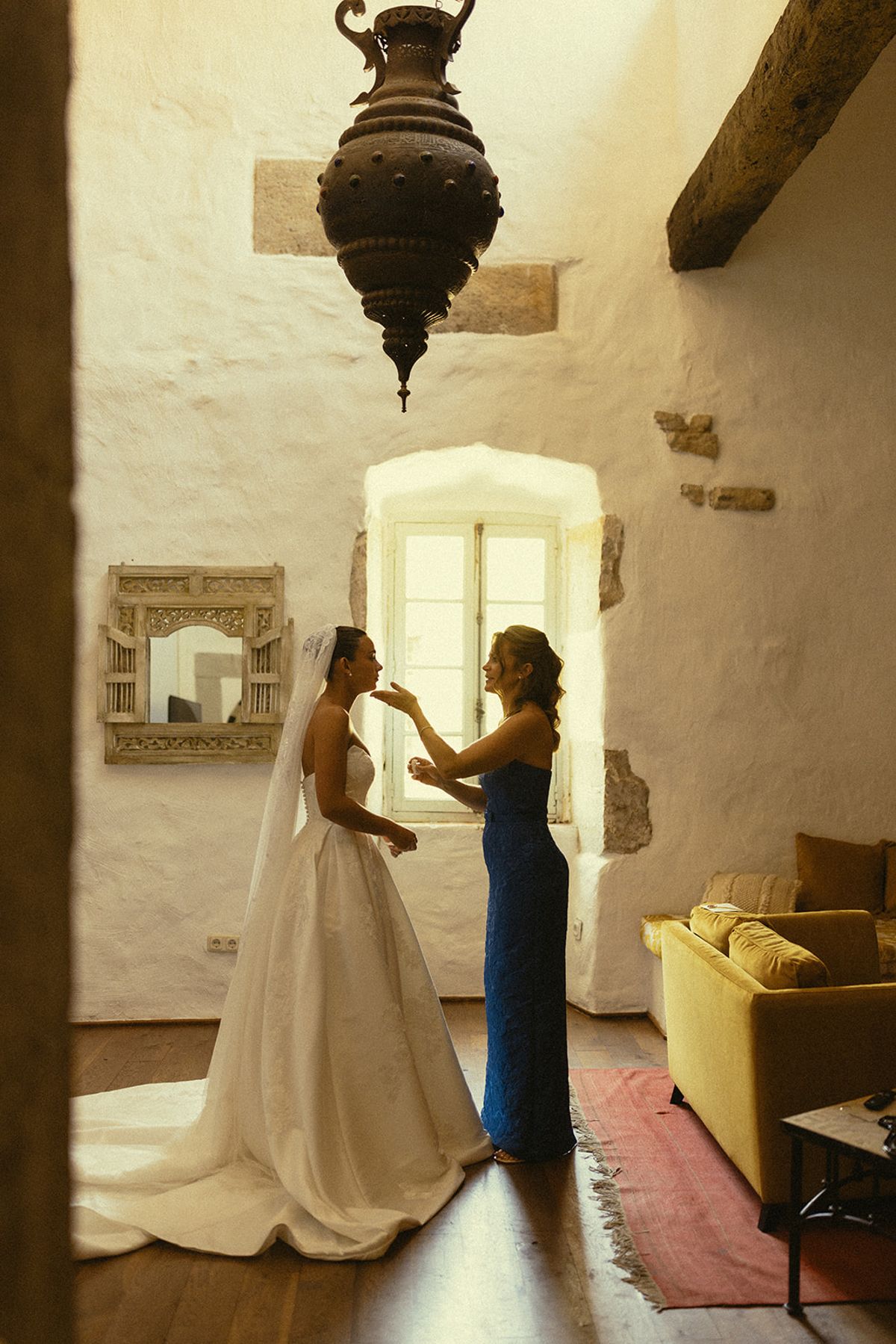 A woman in a blue dress adjusts the veil of a bride in a white gown in a bright stone room.