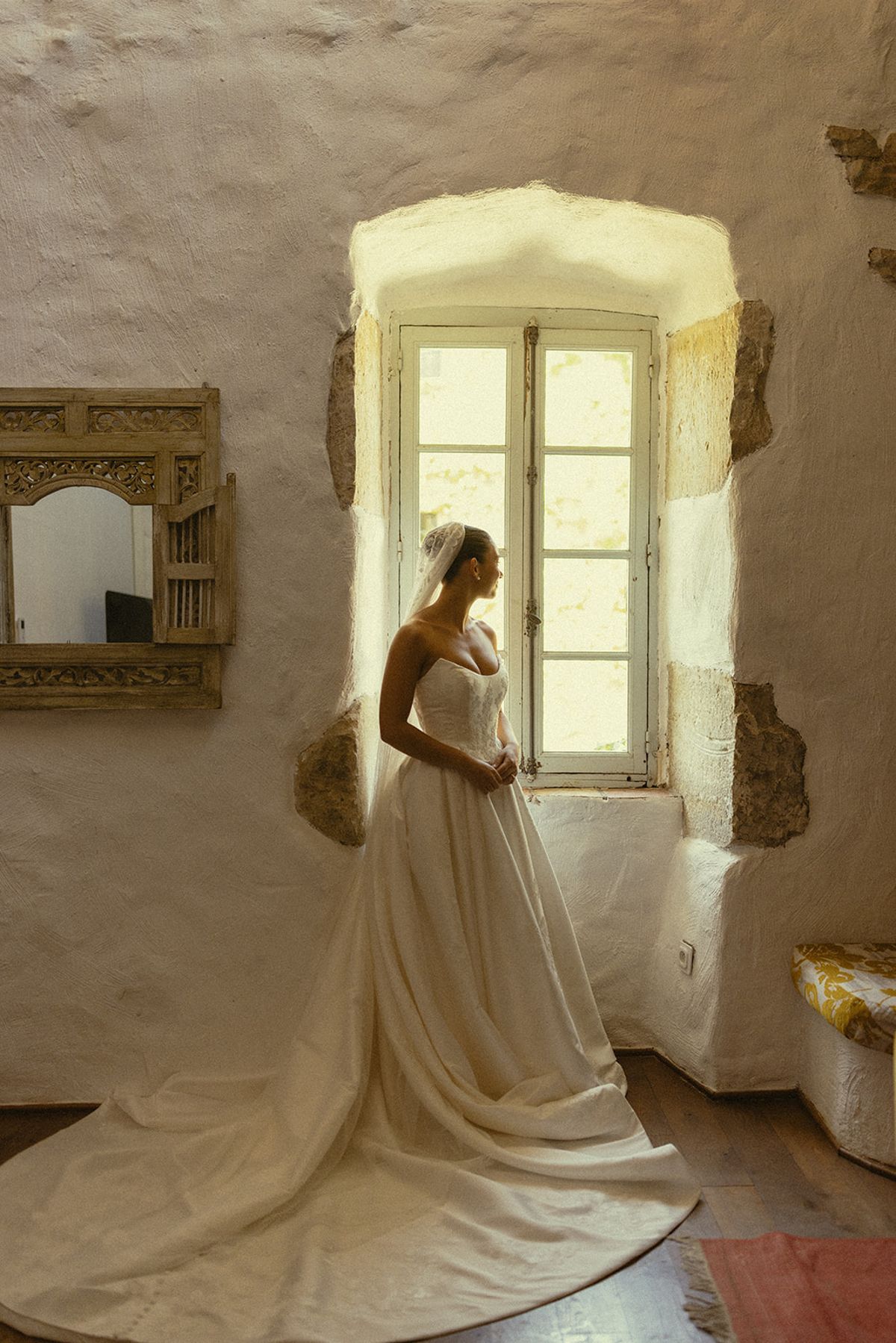 The bride in a long-trained white gown stands in profile beside a tall window in a whitewashed stone room.