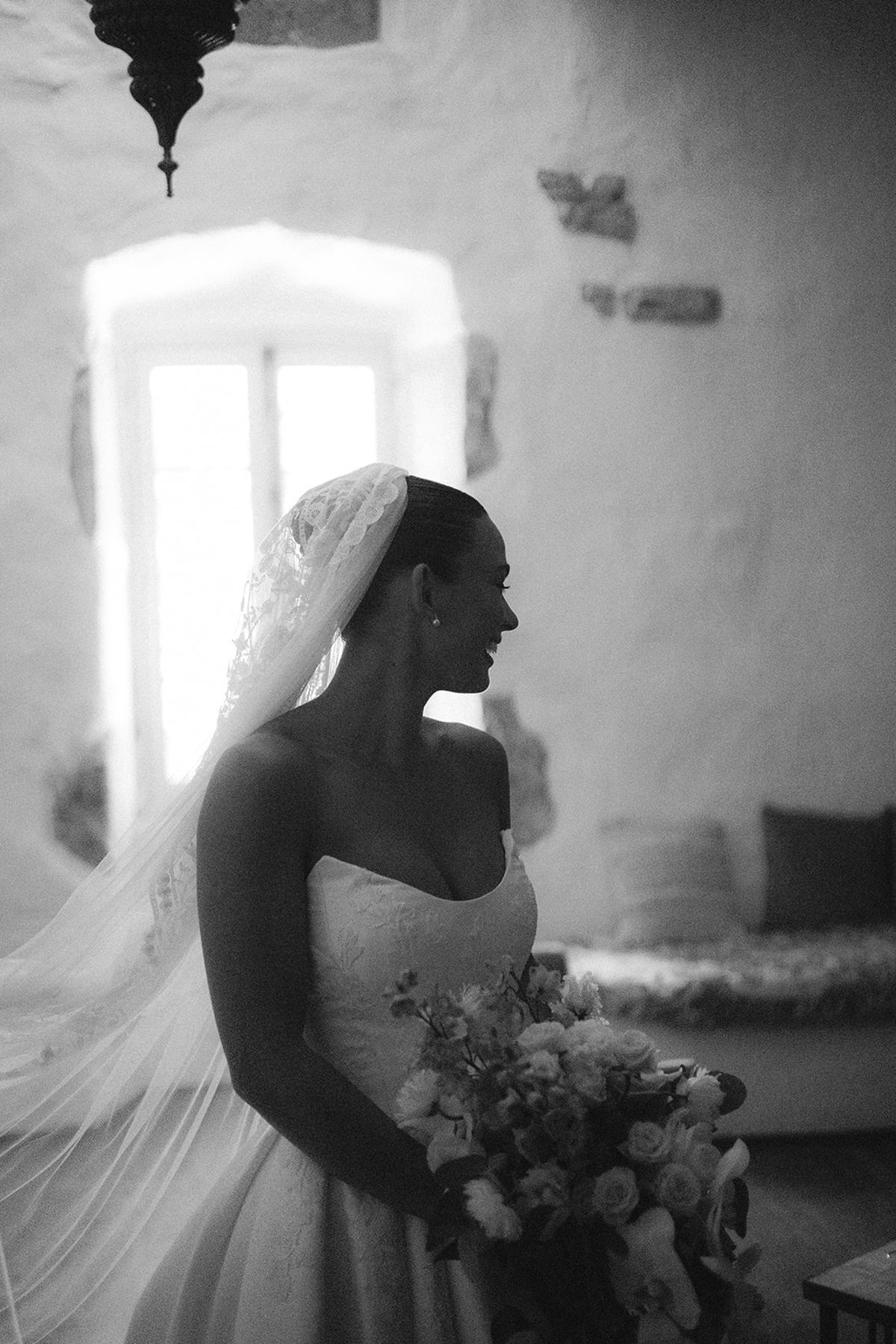 In black and white, a bride in a veil and strapless gown holds a bouquet and looks away beside a bright window.