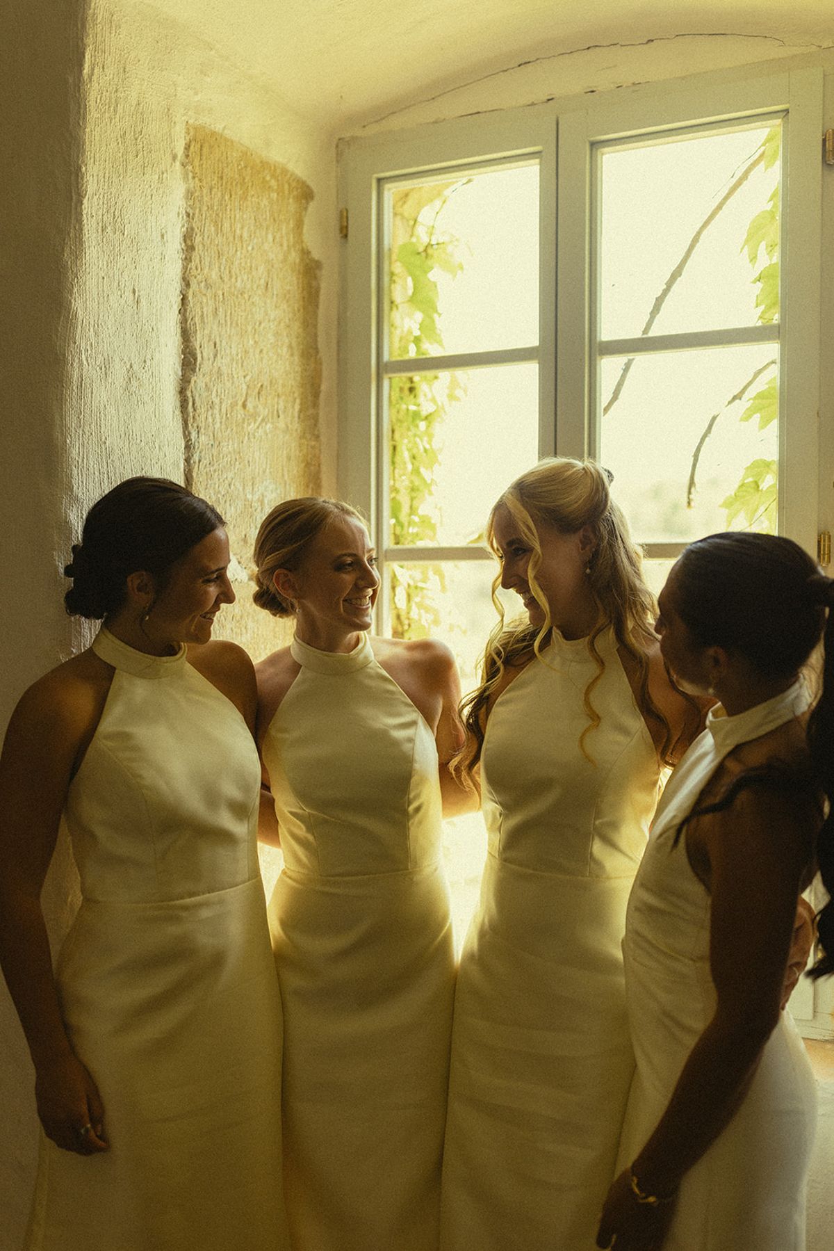 Four bridesmaids in pale yellow halter-neck dresses stand talking together in front of a bright tall window.
