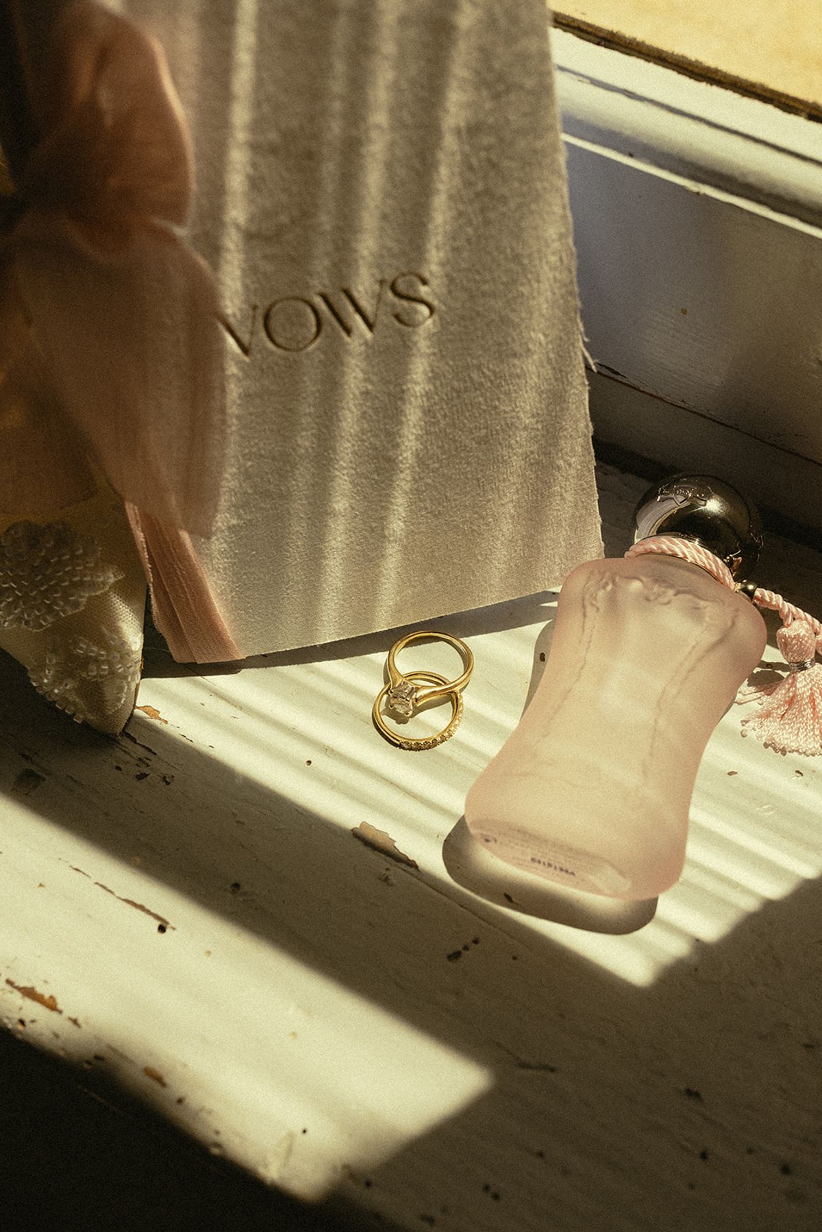 A vows booklet, two gold rings and a pink perfume bottle rest on a windowsill in strong sunlight.