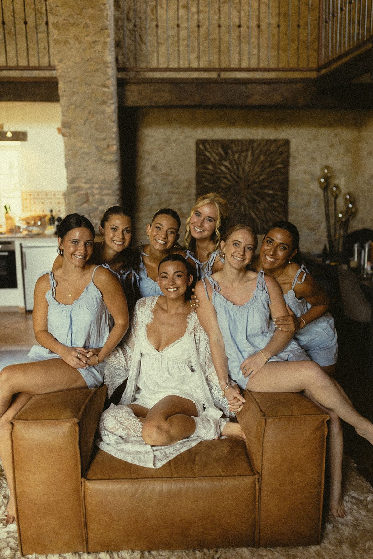 The bride and bridesmaids in matching pale blue robes sit close together on a tan leather sofa, smiling at the camera.