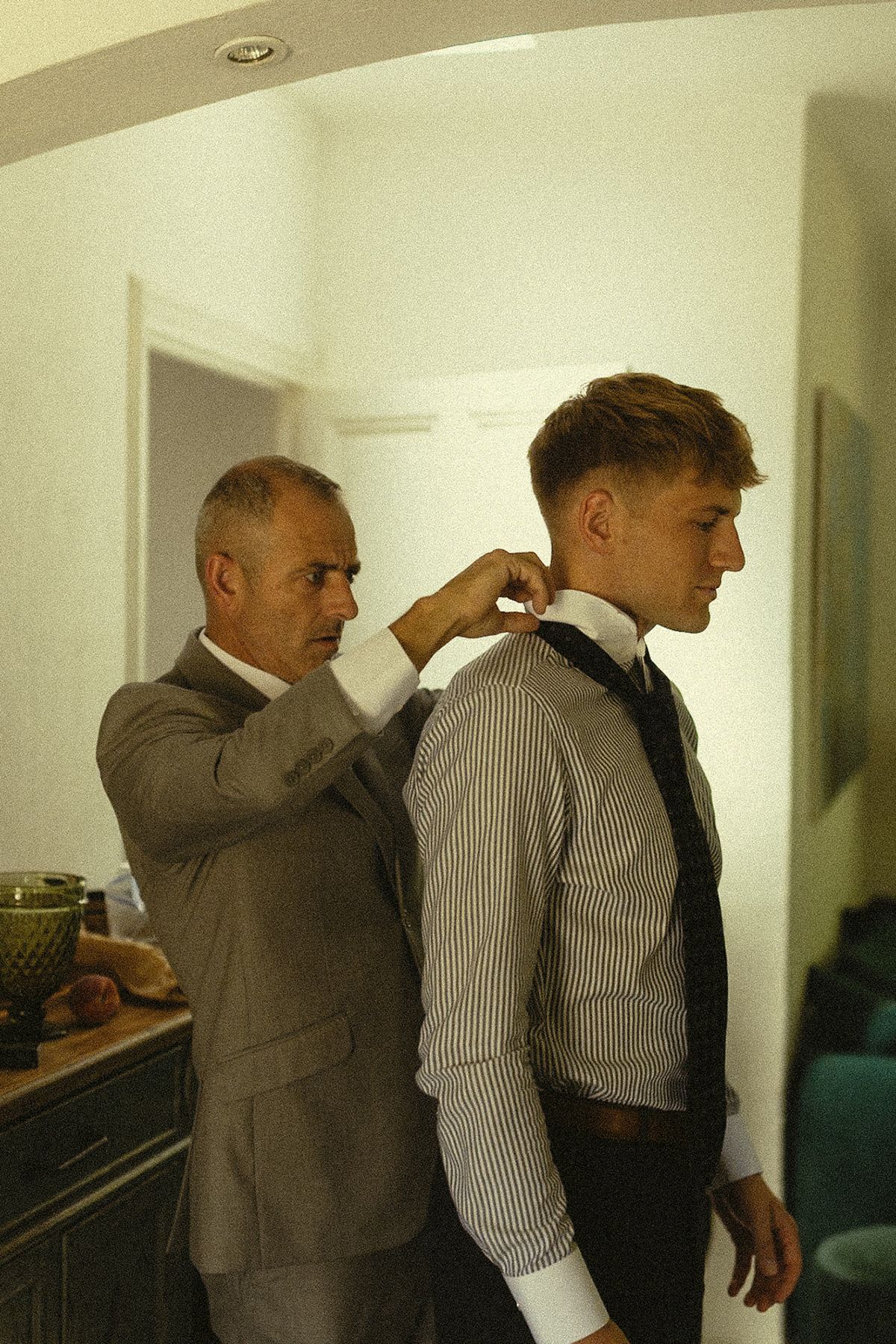 A man in a grey suit adjusts the collar of the groom, who wears a striped shirt and loosely knotted tie, in a bright room.
