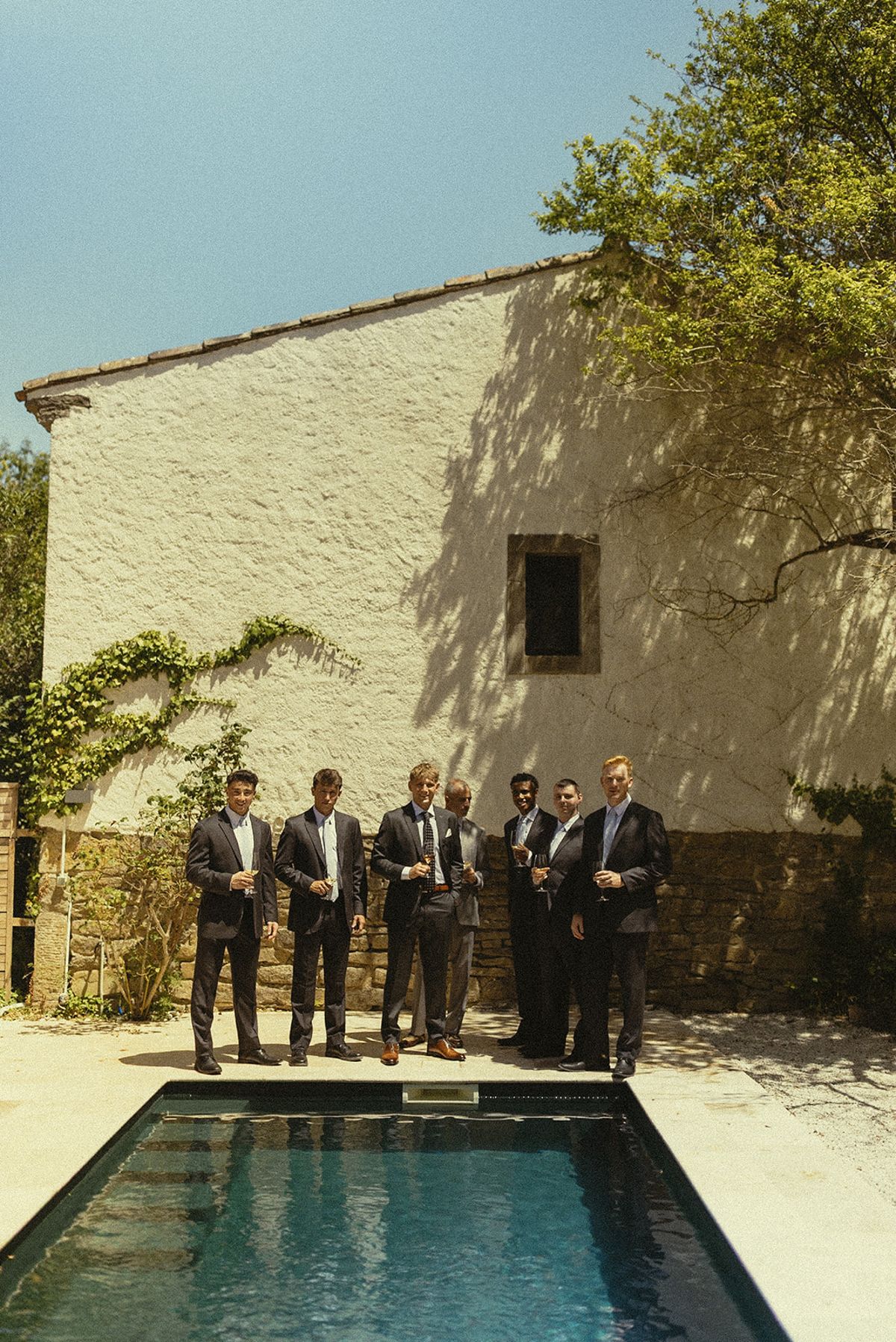 A row of men in dark suits stands beside a small swimming pool, in front of a pale stone building and a tree.