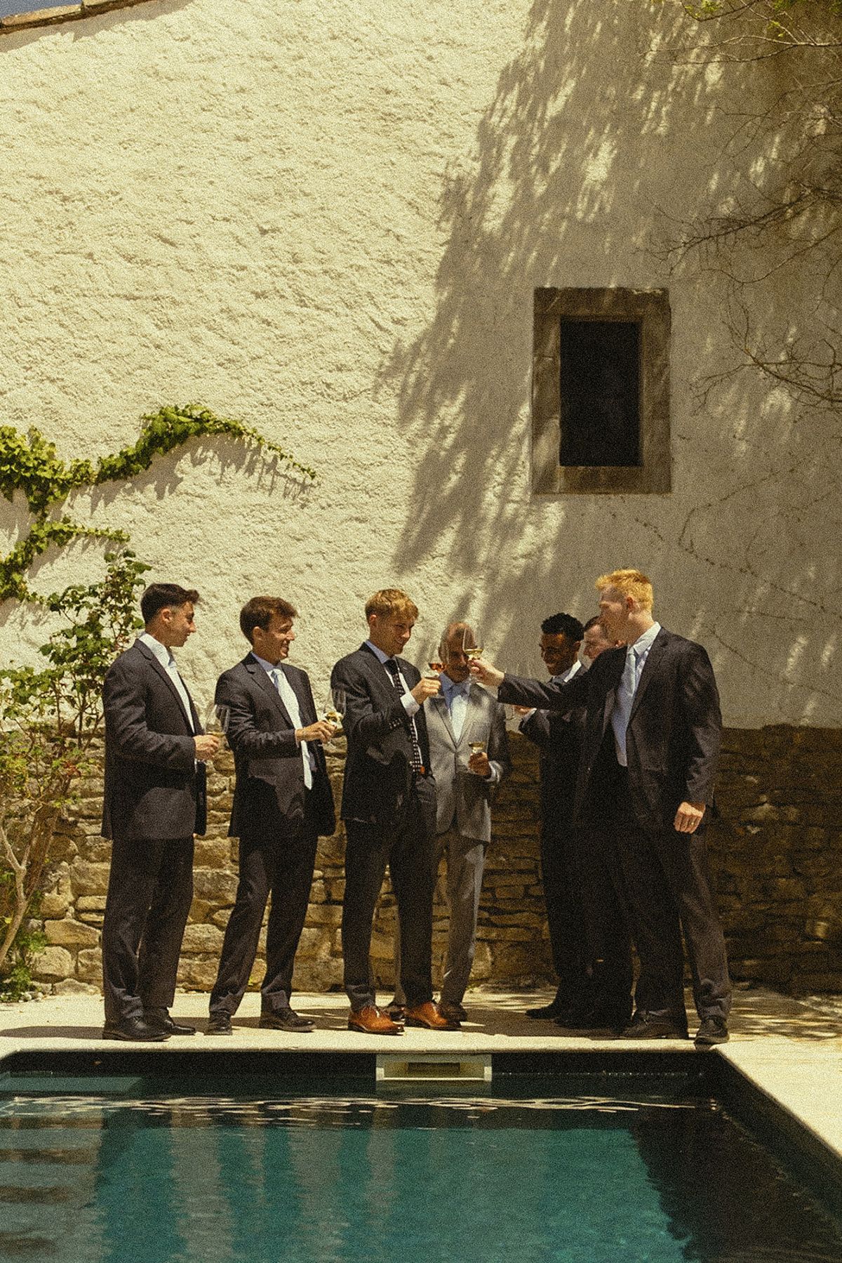 A group of men in dark suits stand talking beside a pool in front of a sunlit whitewashed wall.