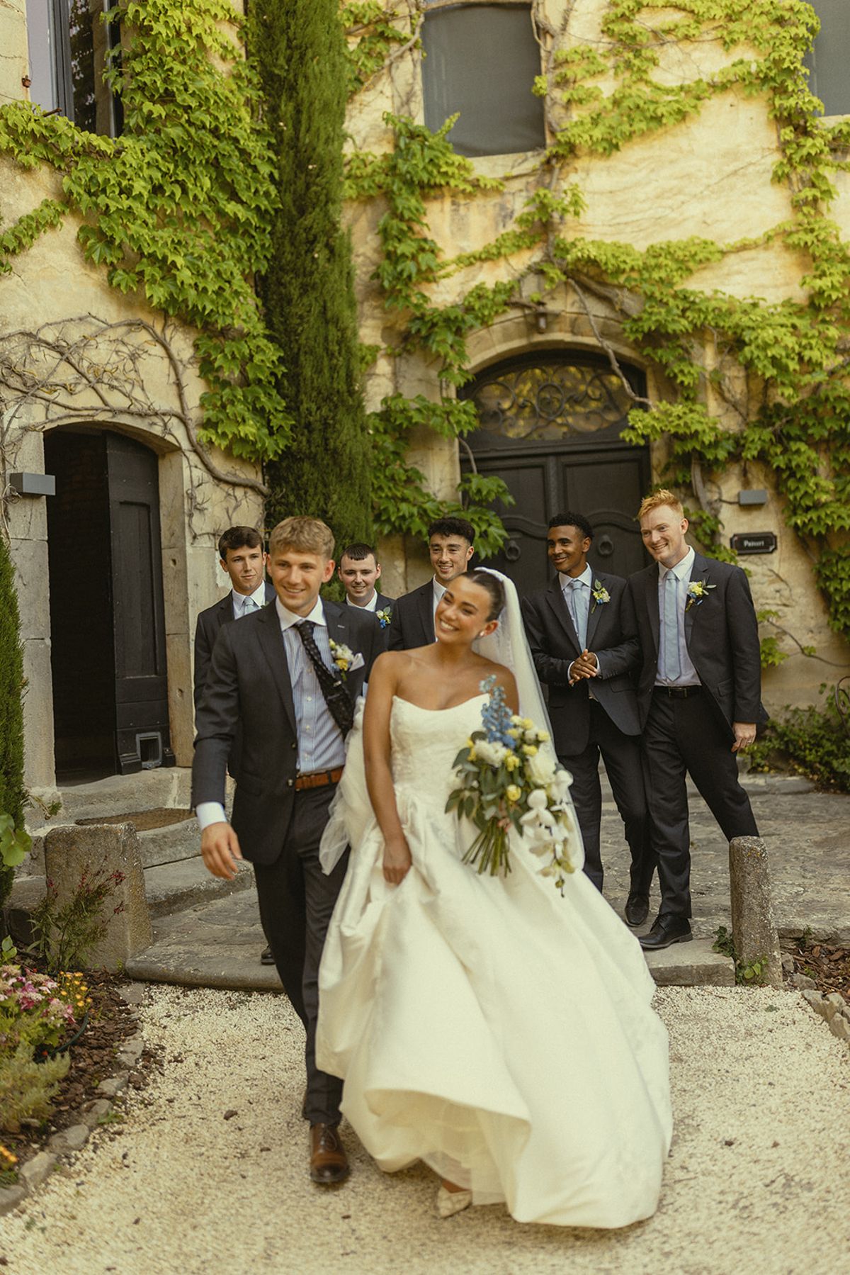 The groom and the laughing bride walk across a gravel courtyard, followed by groomsmen in suits beneath an ivy-covered wall.