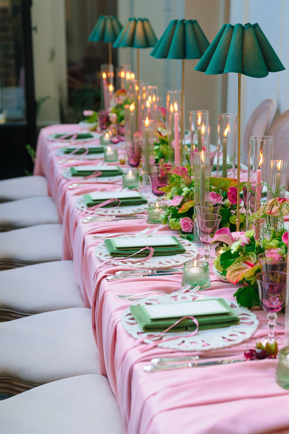 Long dinner table dressed in pink linen with green napkins, taper candles and emerald lamps
