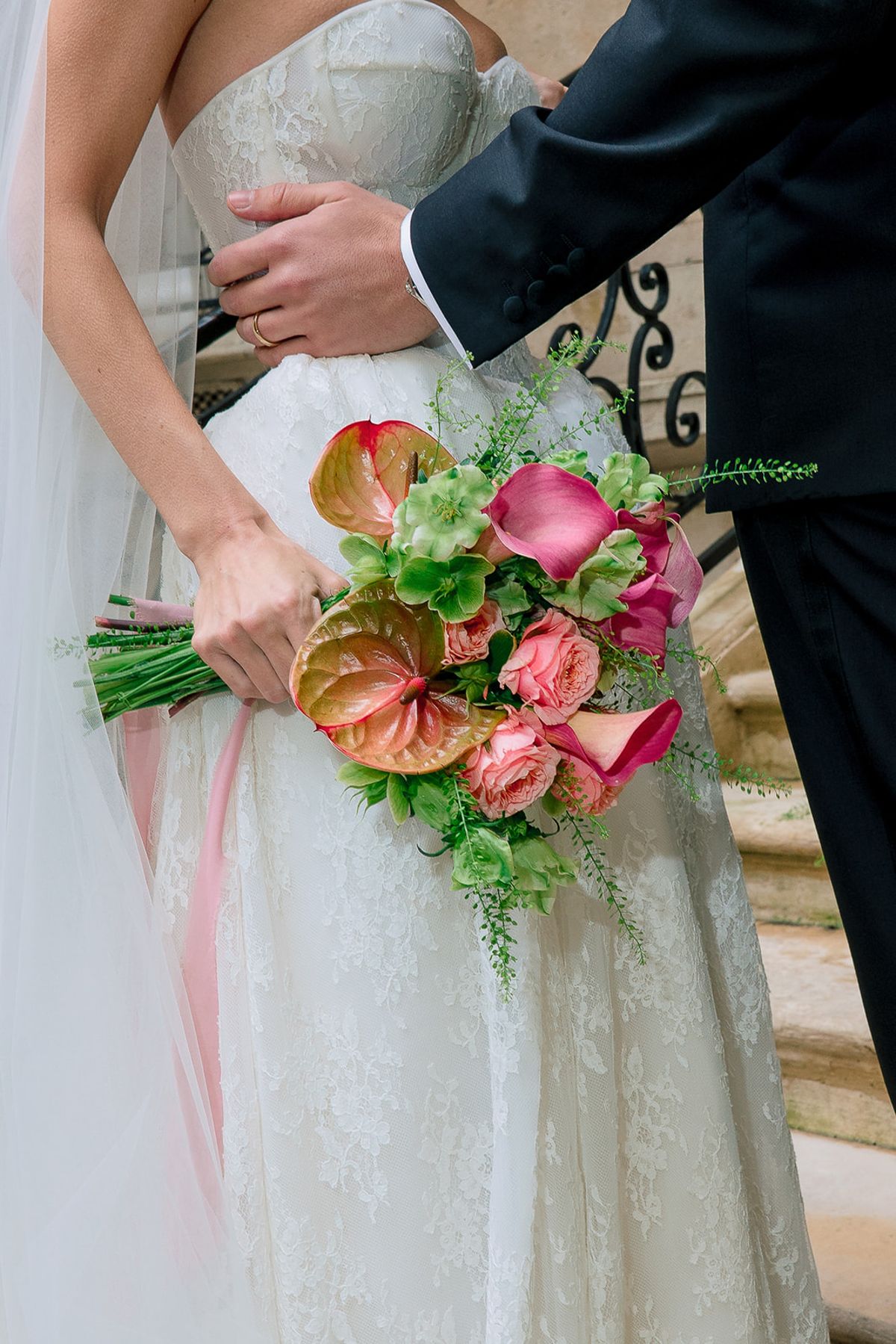Bridal bouquet of pink calla lilies, anthuriums and roses held against a lace gown