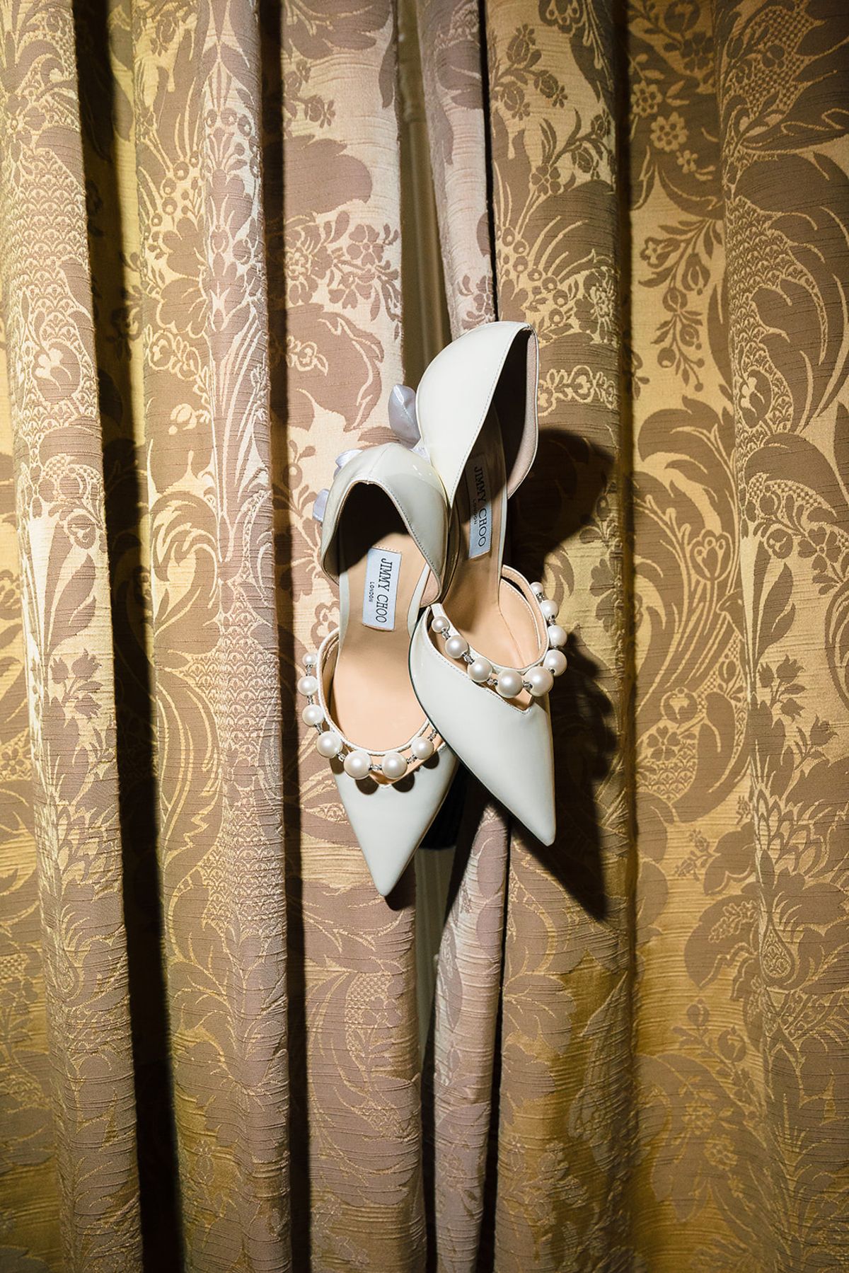 Pearl-embellished bridal heels hanging against a gold damask curtain