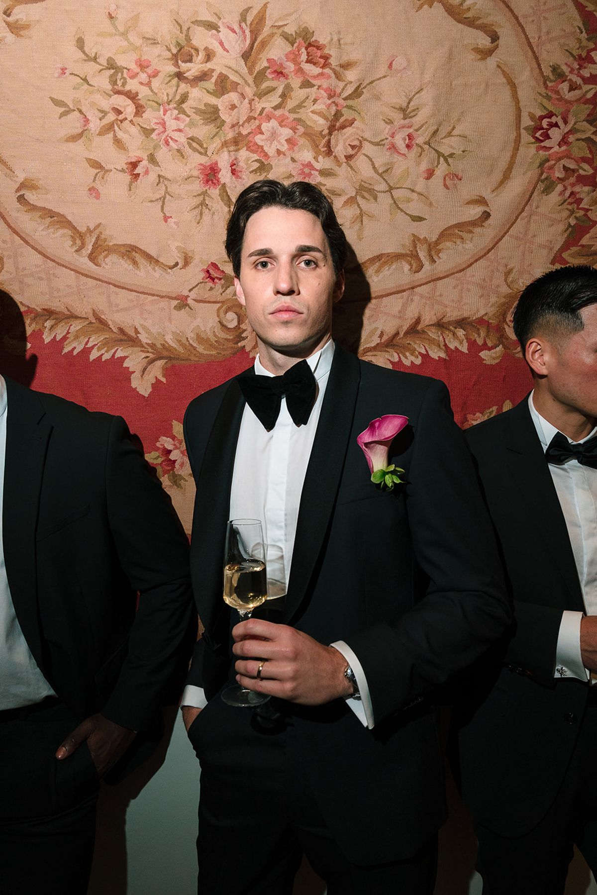 Groom holding a glass of white wine among his groomsmen in front of a floral tapestry
