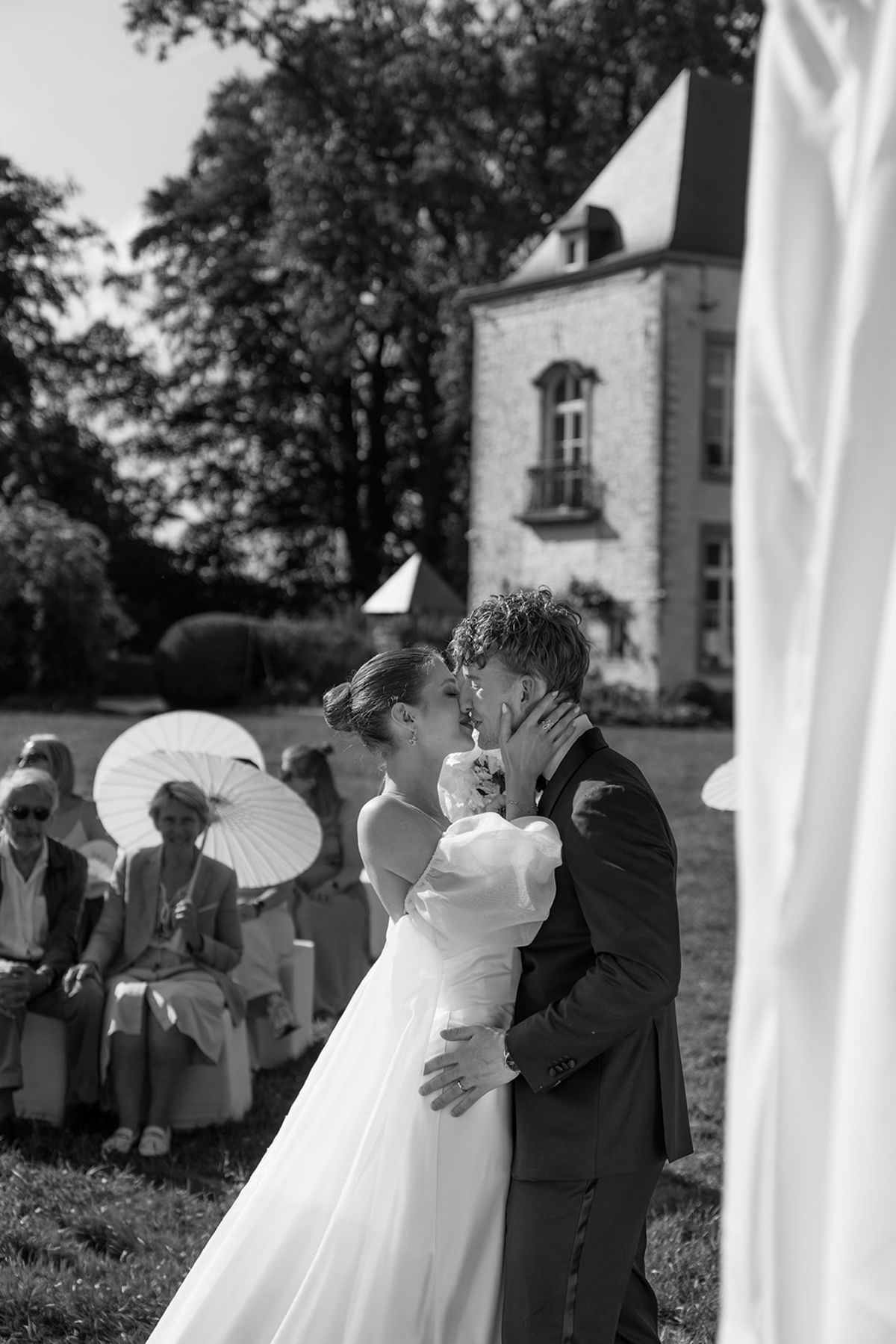 Bride and groom close together at the ceremony, guests with parasols beyond, black and white
