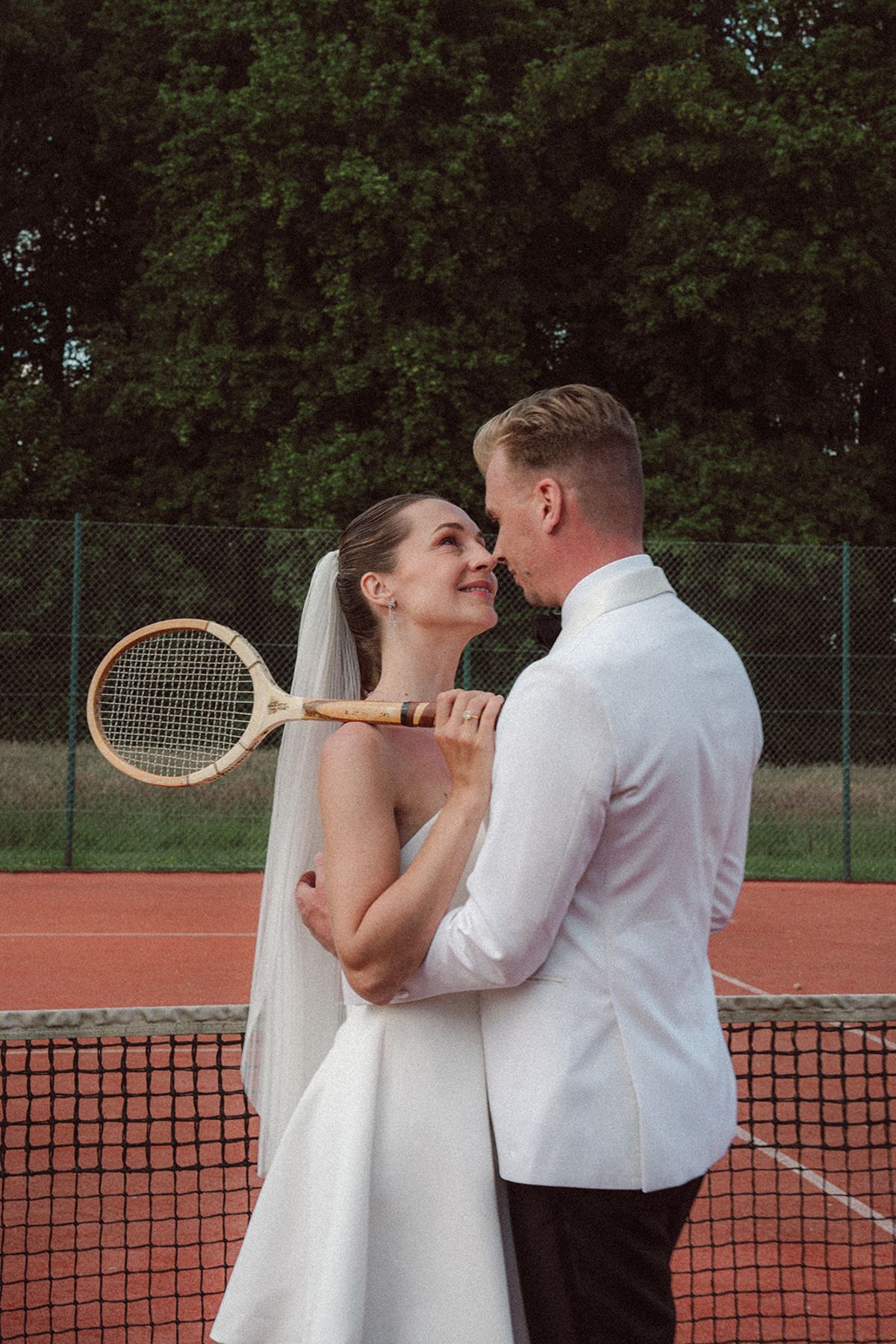 Bride and groom embracing on the tennis court, groom holding a racket