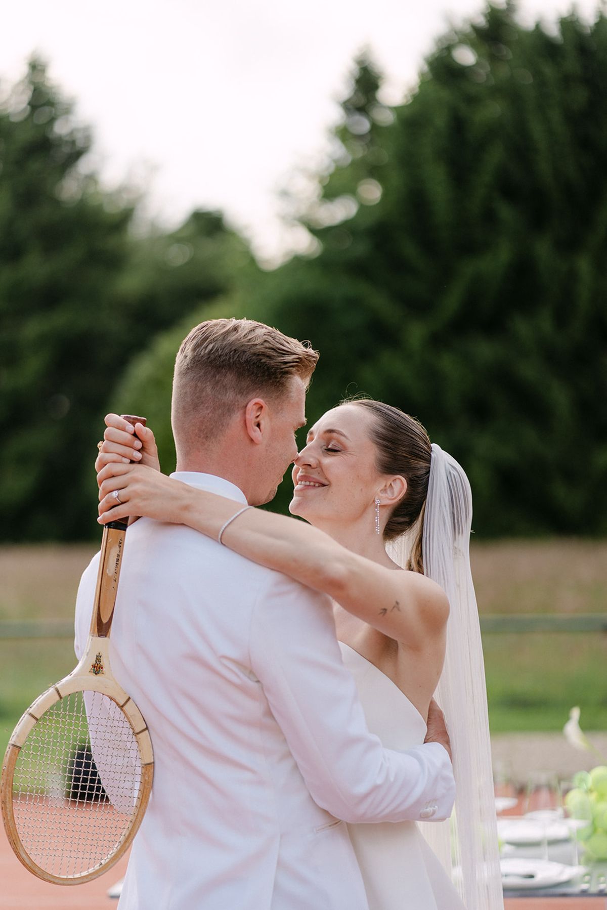 Bride and groom embracing on the tennis court, groom holding a racket
