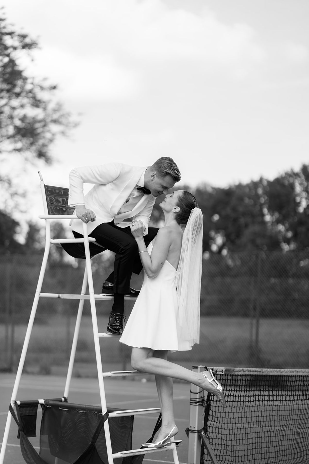 Groom kneeling to kiss the bride, seated on the tennis umpire's chair