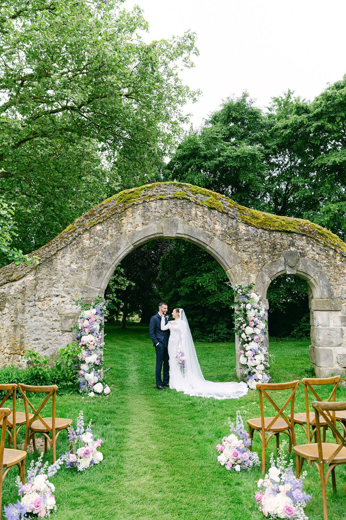 The couple stand far off on a grass aisle beneath a mossy stone arch, with wooden chairs and flowers lining both sides and trees behind.