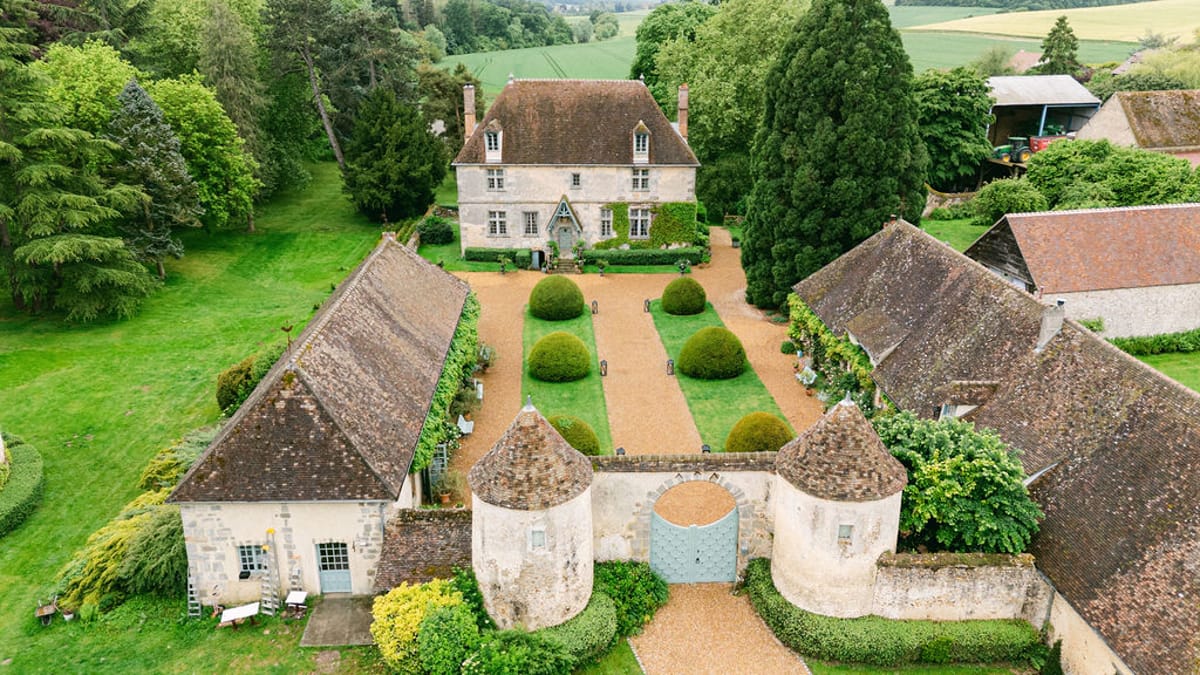 An aerial view of a stone chateau with a tiled roof, a formal garden with clipped hedges, and surrounding outbuildings, trees and fields.